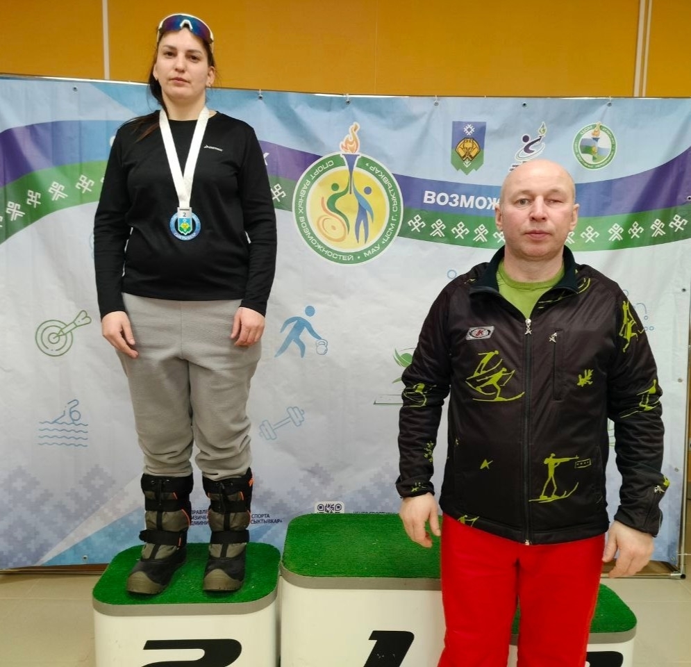 Женщина с медалью на подиуме и мужчина в спортивной куртке рядом, на фоне баннера.
