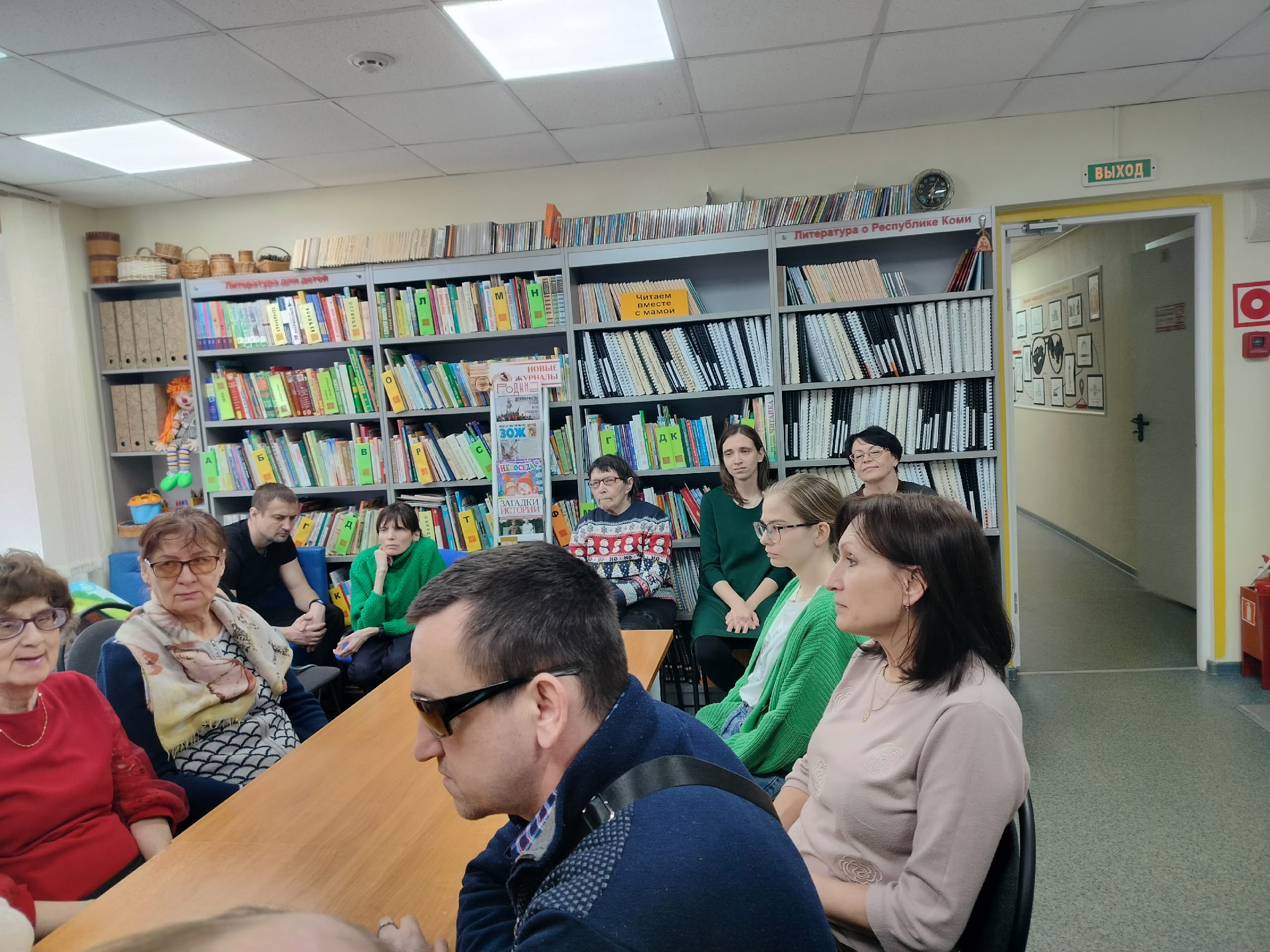 Группа людей сидит за длинным столом в библиотеке, слушая презентацию. На заднем плане видны книжные полки.