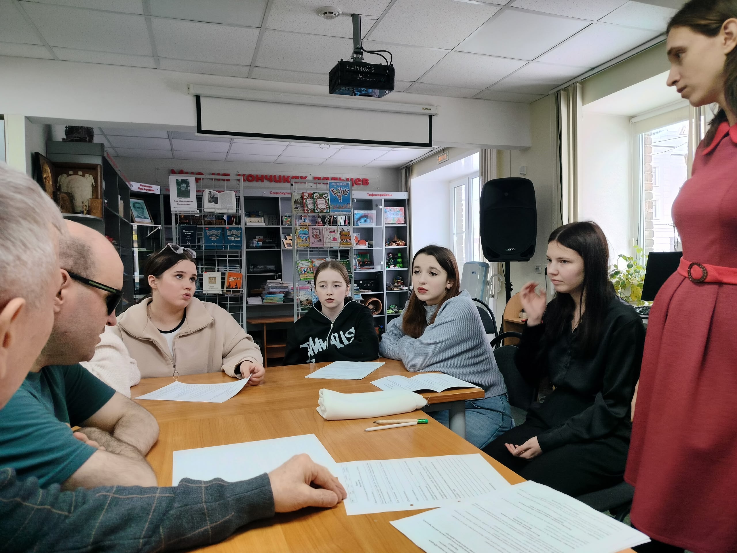 Группа людей сидит за столом с бумагами, внимательно слушая, на фоне книжные полки и проектор.