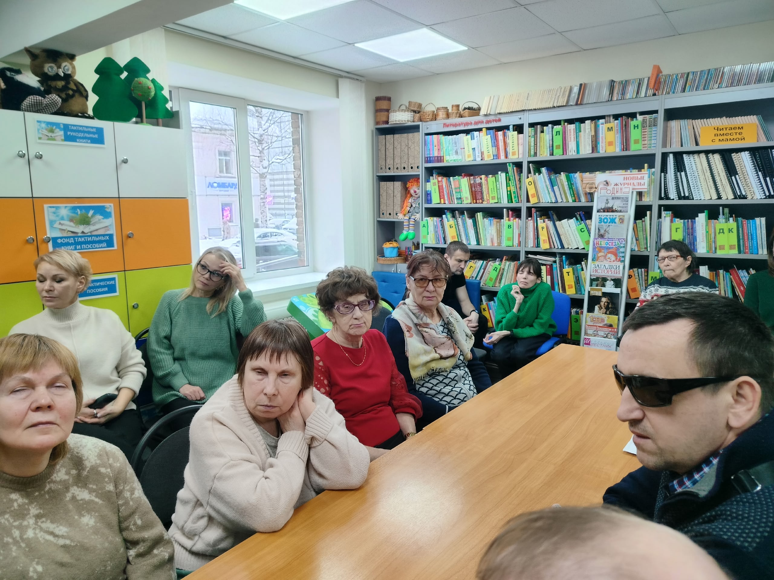 Участники мероприятия сидят за столом в библиотеке, внимательно слушая. Мужчина в очках справа.