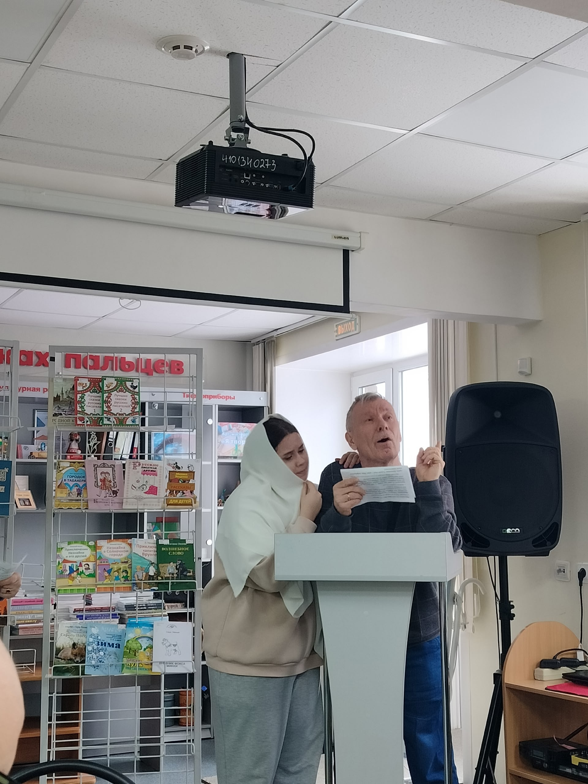 Мужчина выступает с трибуны, рядом стоит девушка в платке, на фоне книжные стеллажи и проектор.