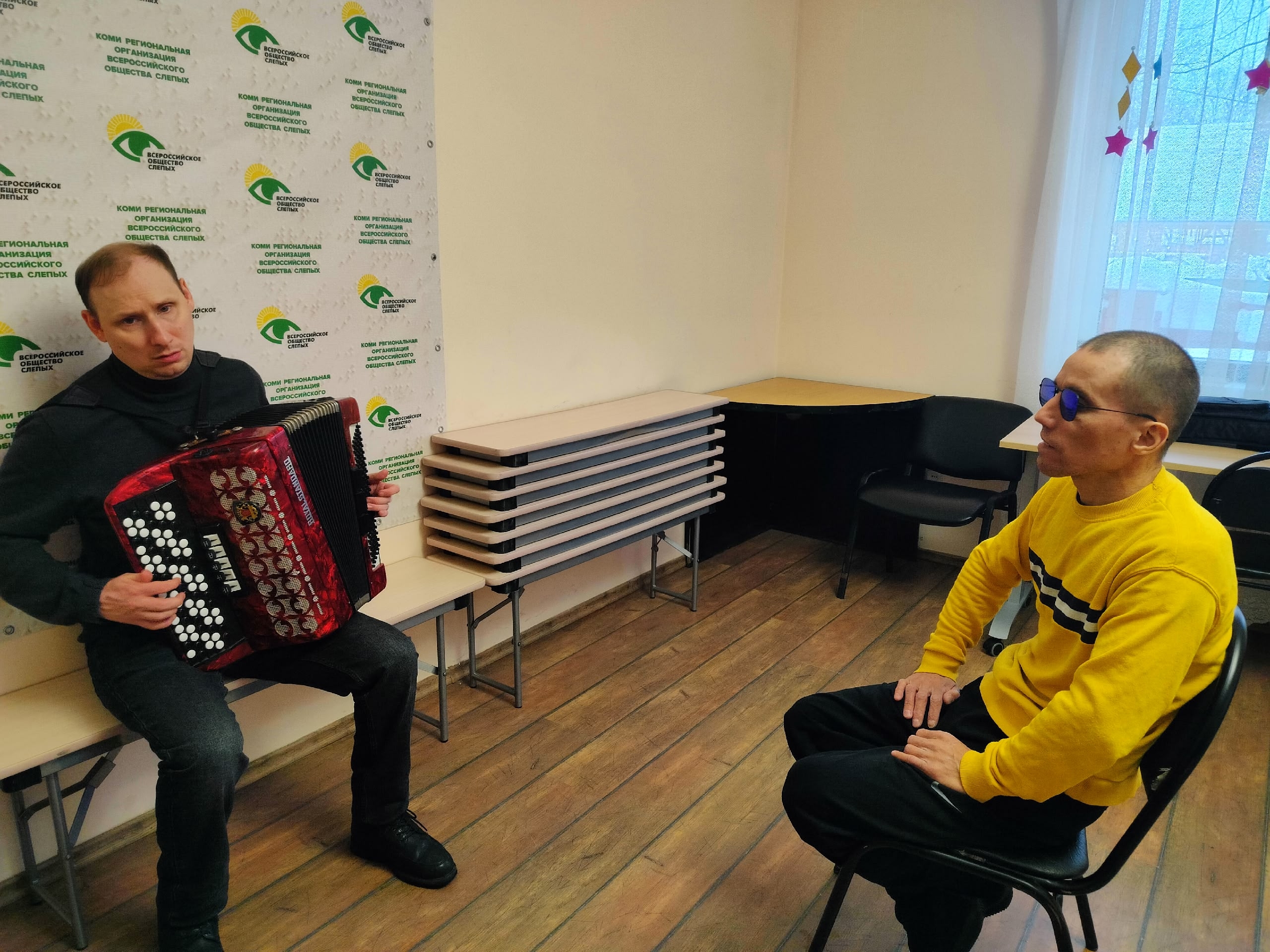 Мужчина в черном играет на красном аккордеоне, а другой мужчина в желтом свитере и синих очках слушает его в комнате с логотипами.