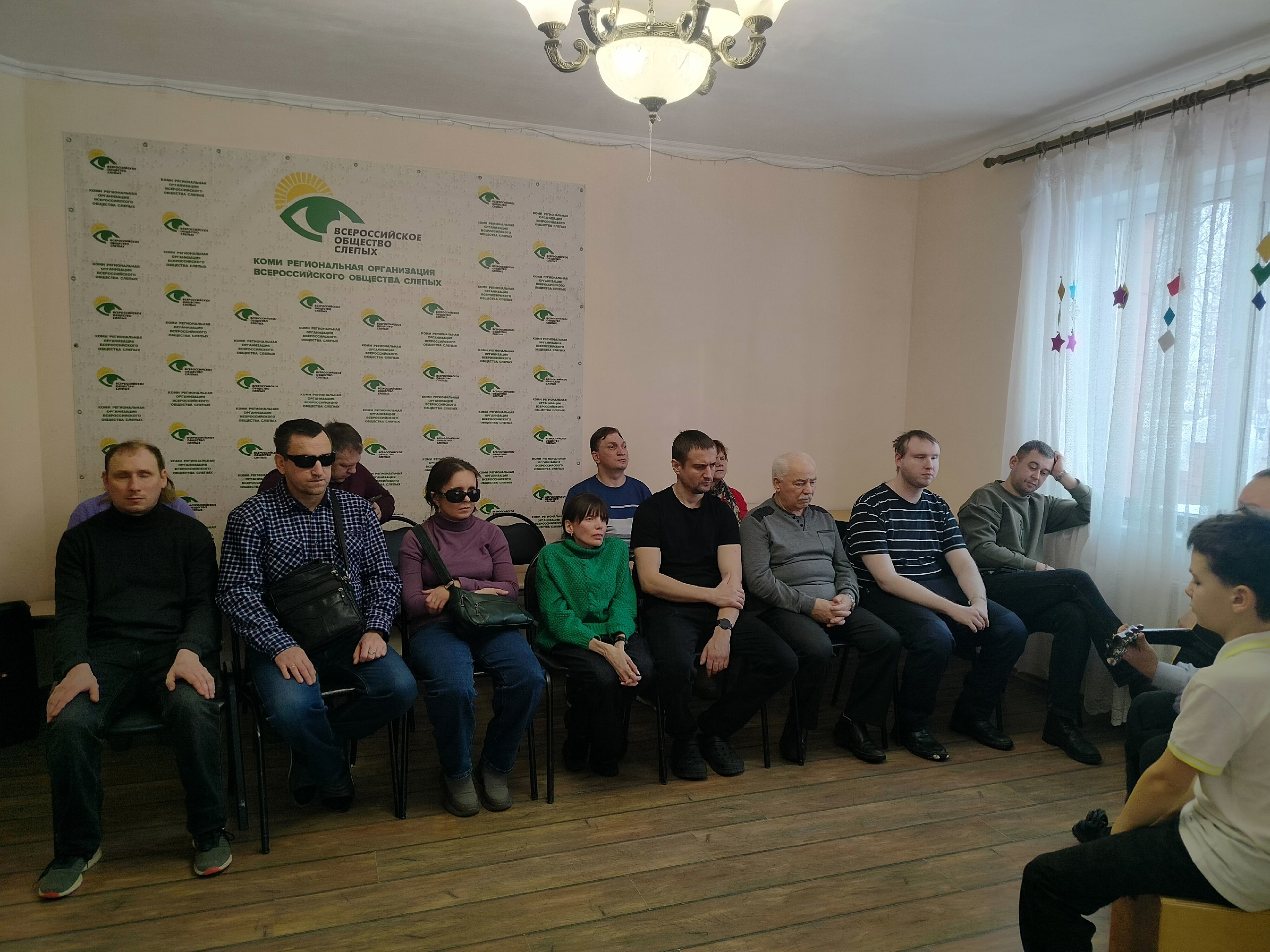 Группа людей, включая незрячих, сидит в зале с баннером Всероссийского Общества Слепых.
