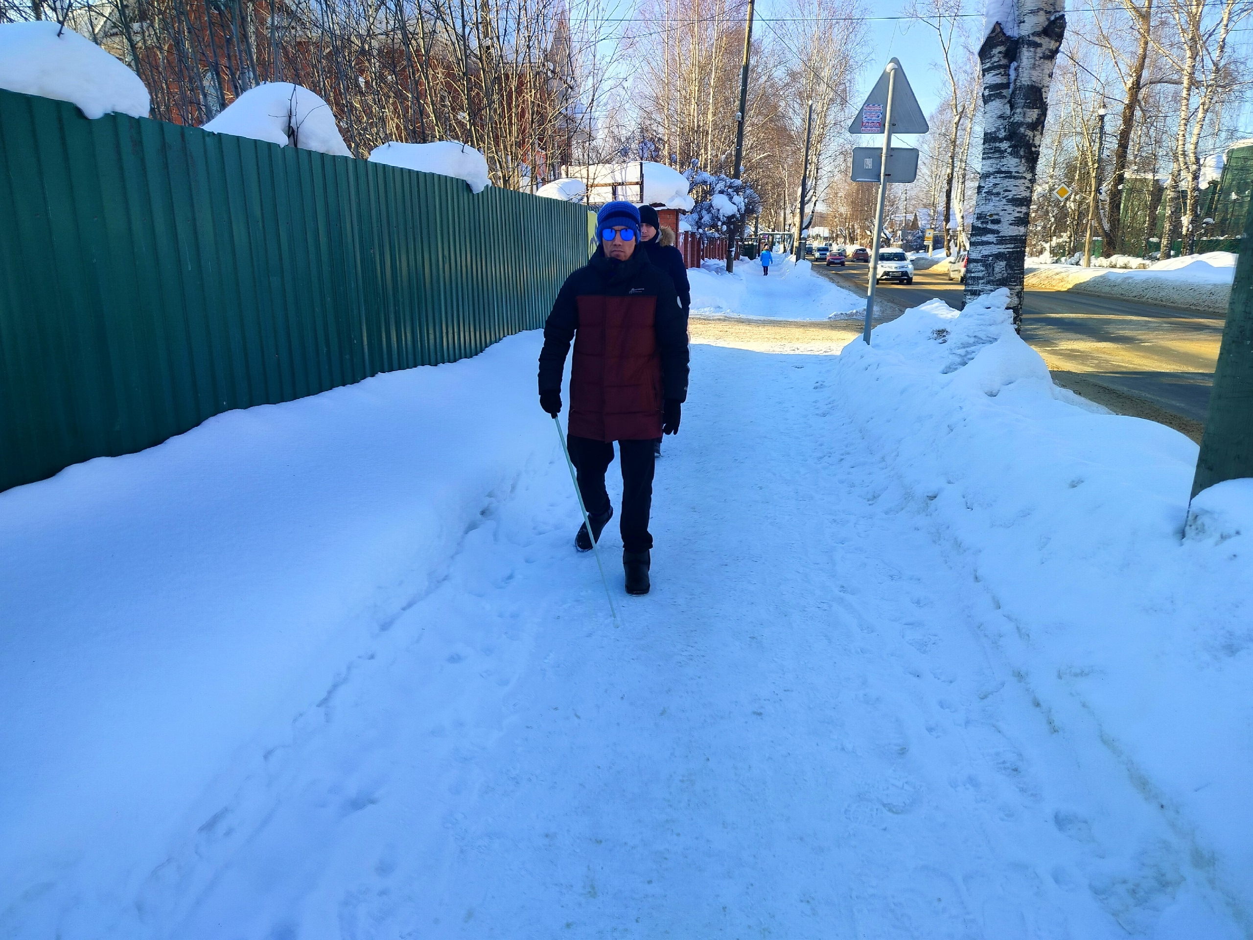 Мужчина с белой тростью идет по заснеженному тротуару вдоль зеленого забора, тренируя ориентировку.