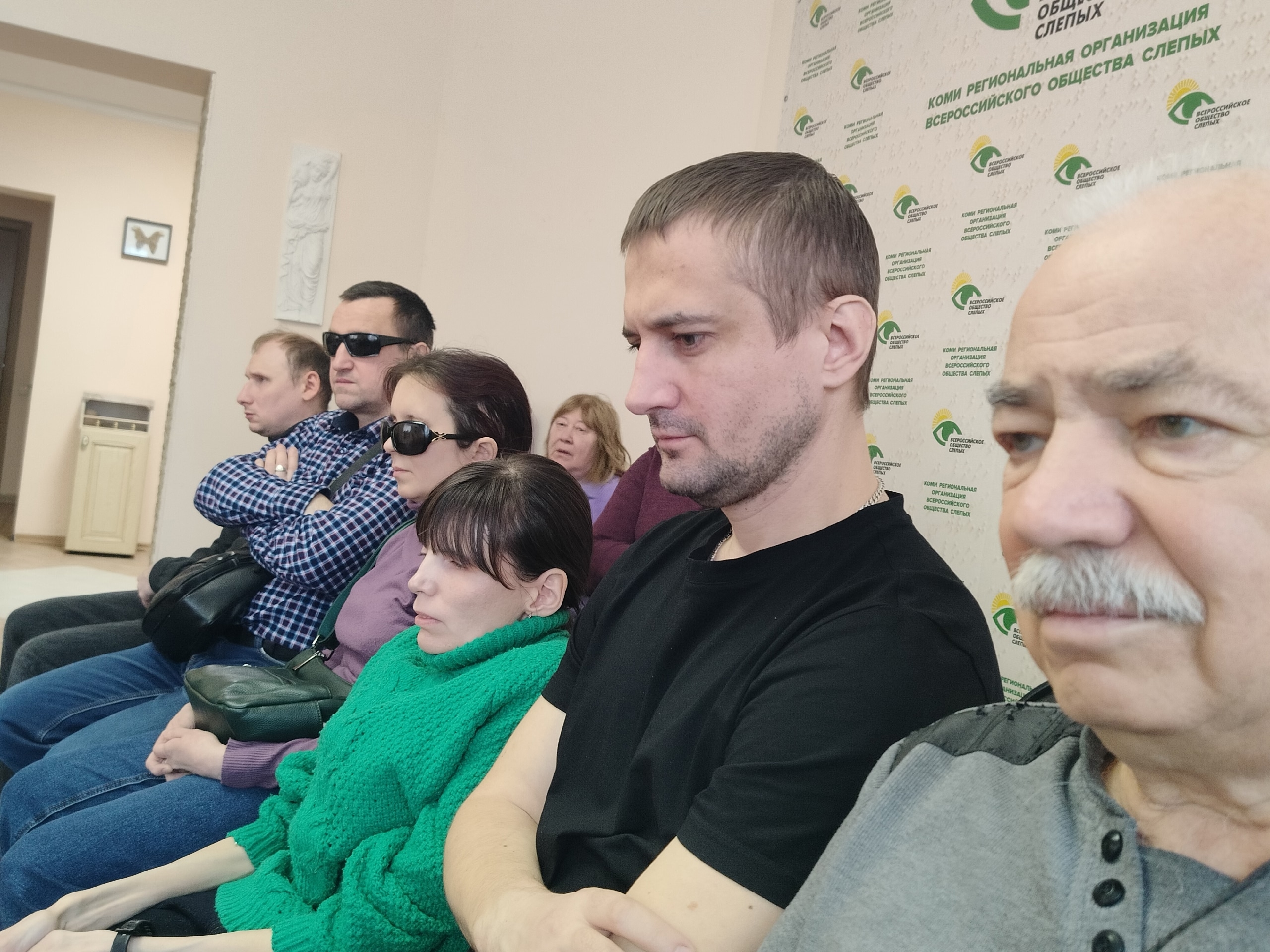 Крупный план слушателей, сидящих в зале, некоторые из них в темных очках.