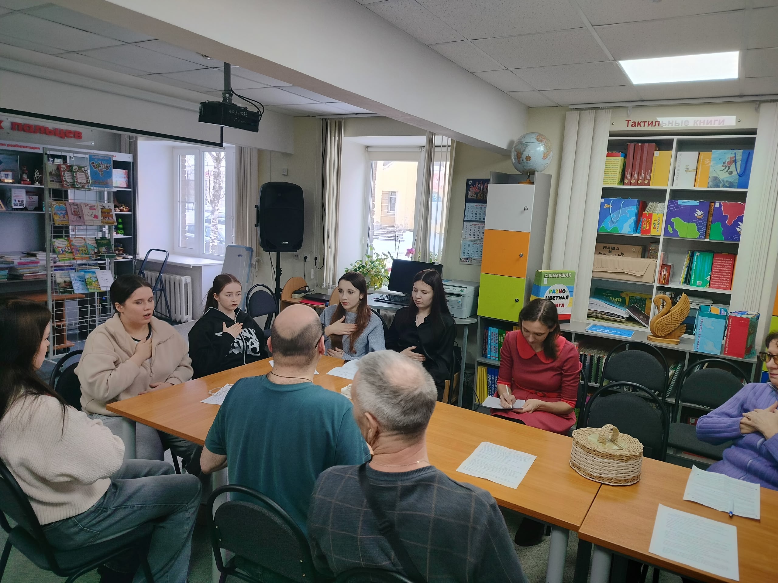 Люди сидят за столом в библиотеке, слушают друг друга, на столе лежат бумаги, вокруг книжные стеллажи.