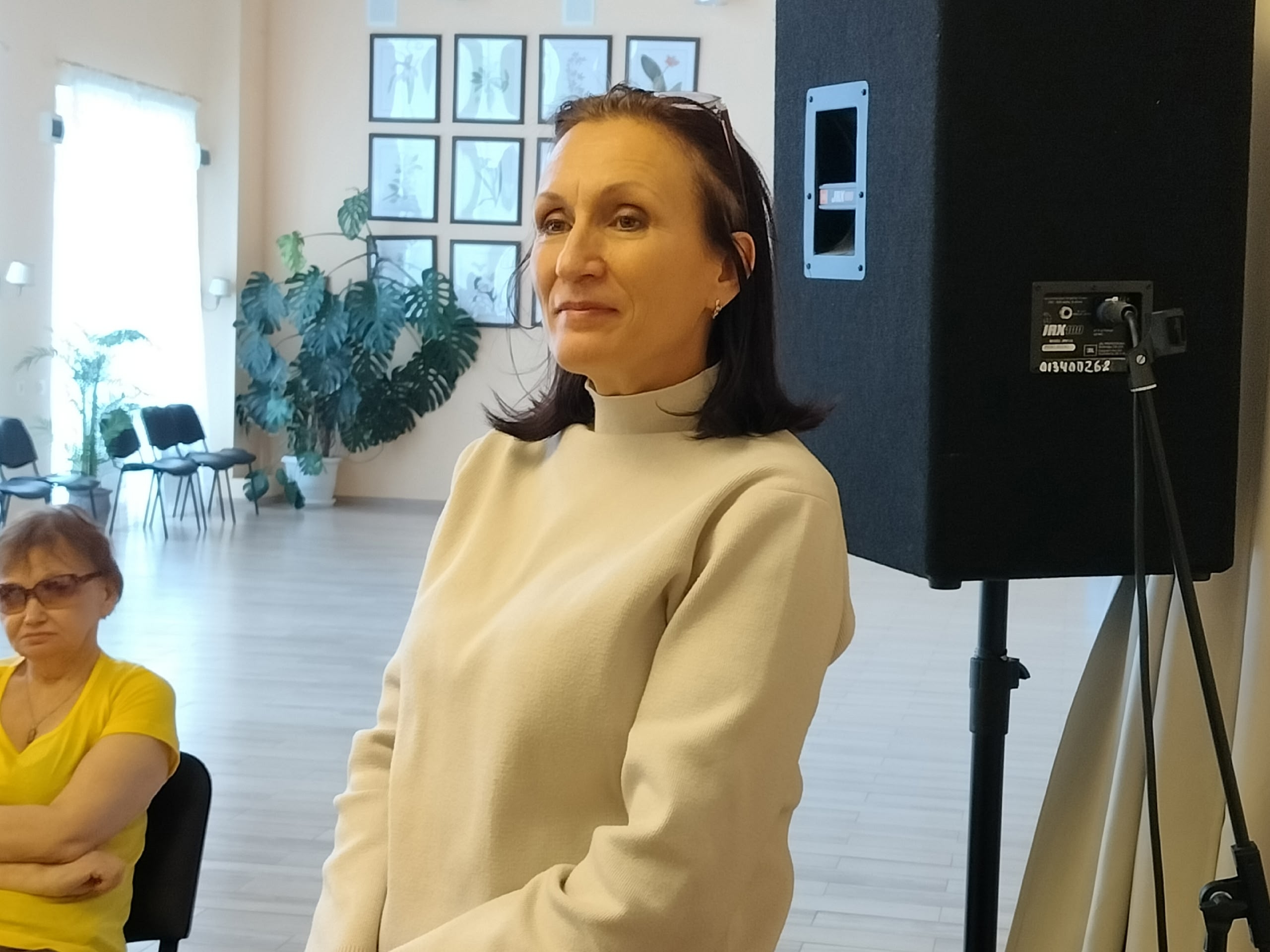 Женщина в светлом свитере стоит, внимательно слушая, на мероприятии.
