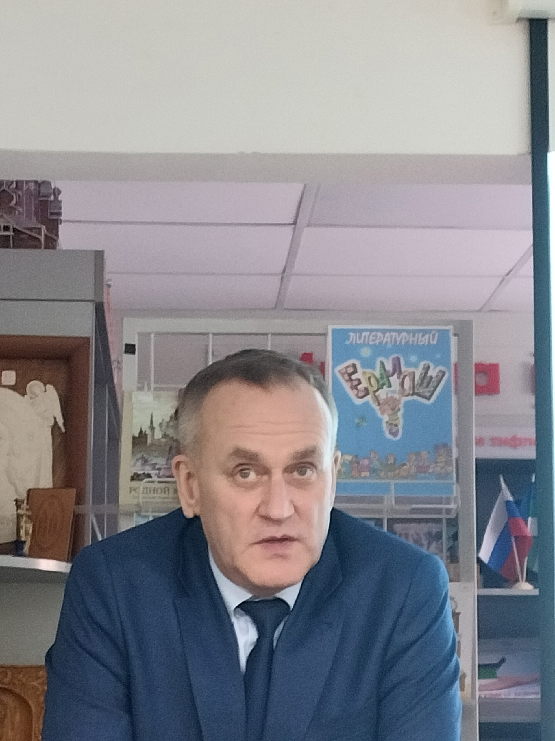 Мужчина в темно-синем костюме и галстуке смотрит вперед, находясь в помещении библиотеки.