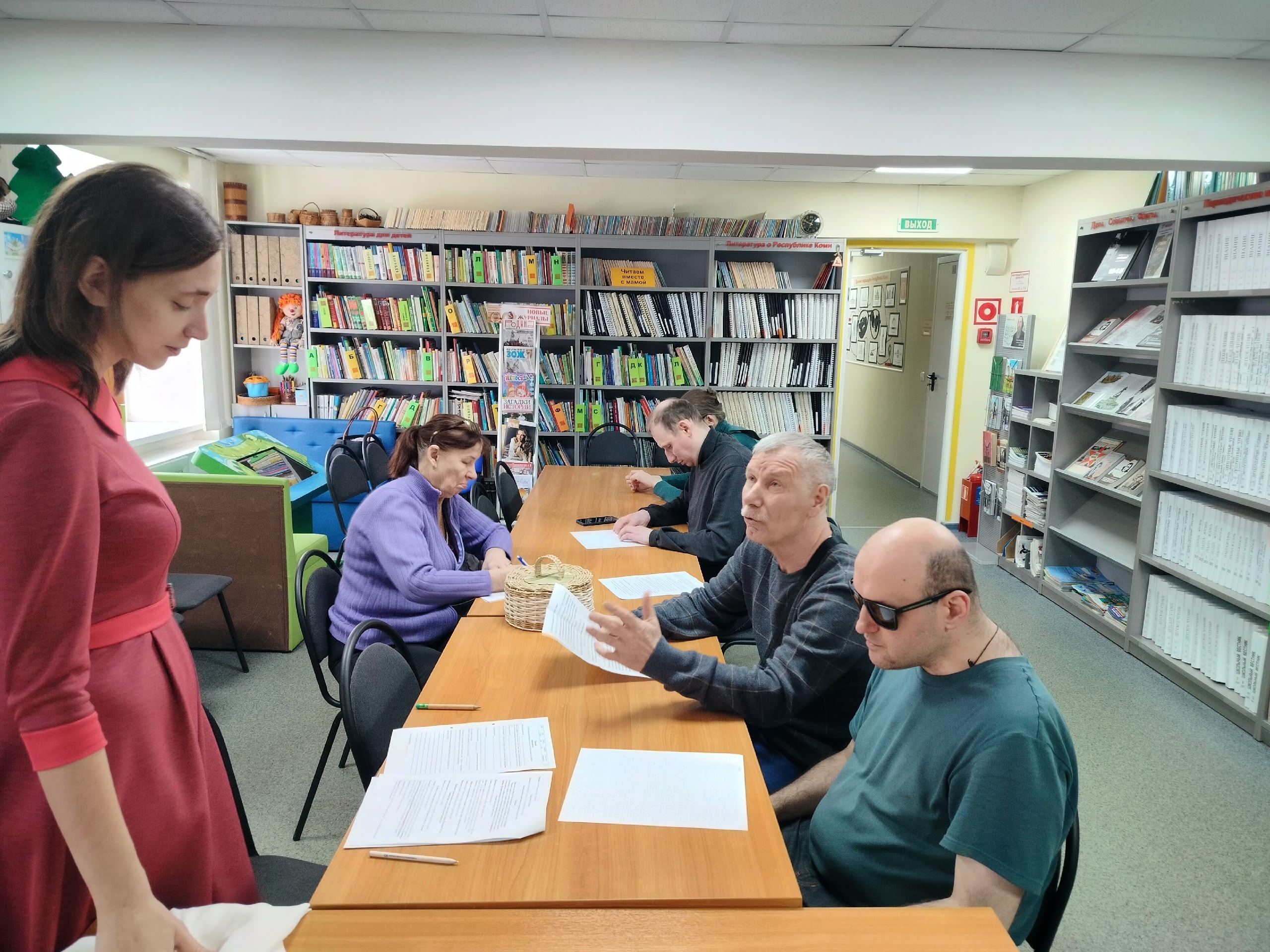 Женщина в красном платье стоит, наблюдая за людьми, сидящими за столами с бумагами в библиотеке.