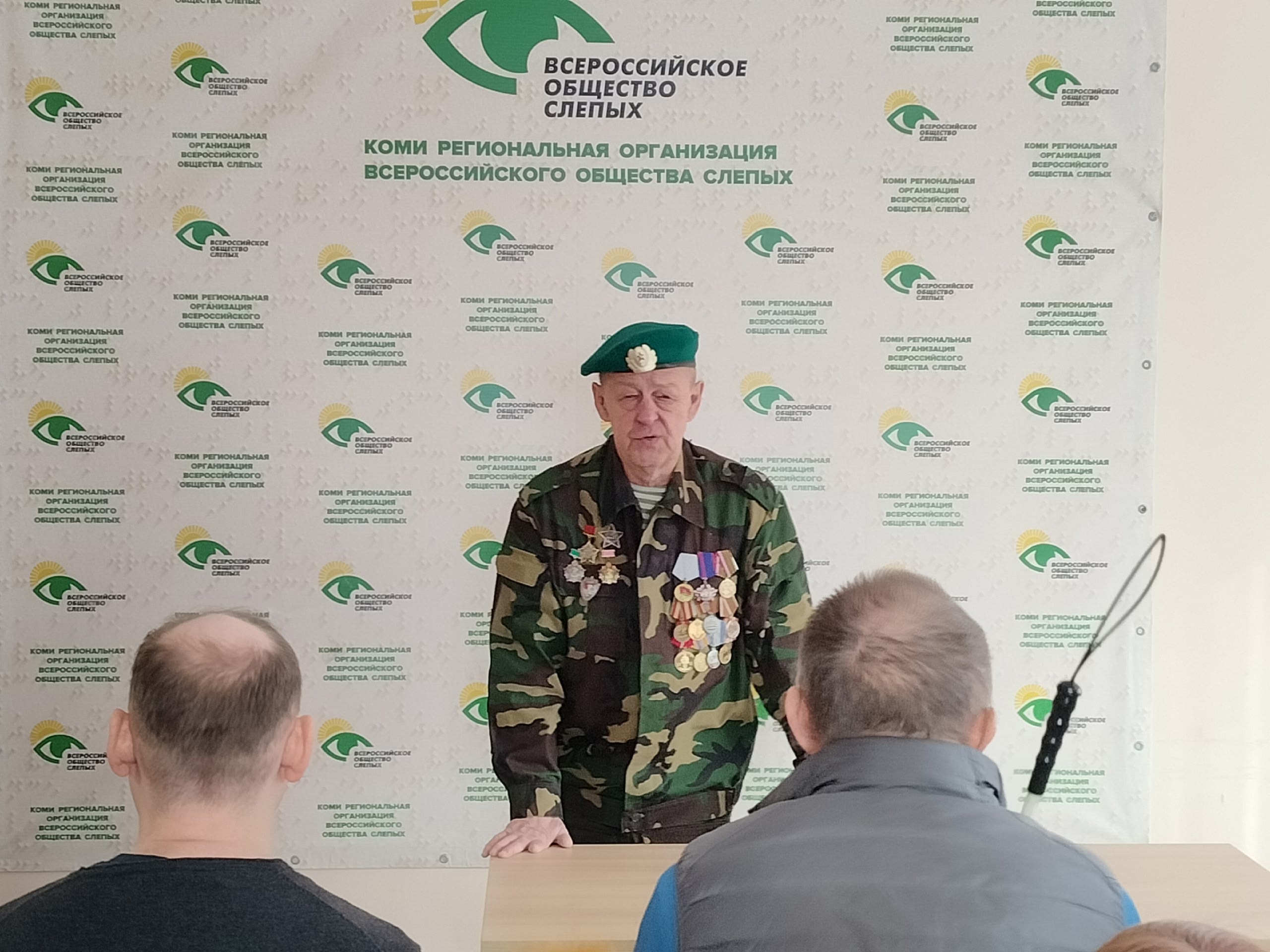Ветеран в камуфляжной форме с медалями и зеленым беретом выступает перед аудиторией на фоне логотипа ВОС.