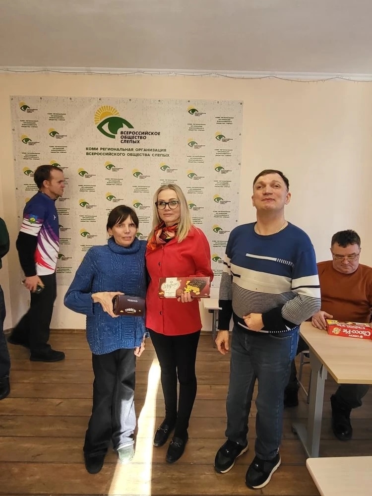 Две женщины и мужчина стоят в помещении, женщины держат в руках подарочные коробки, на заднем плане баннер организации.
