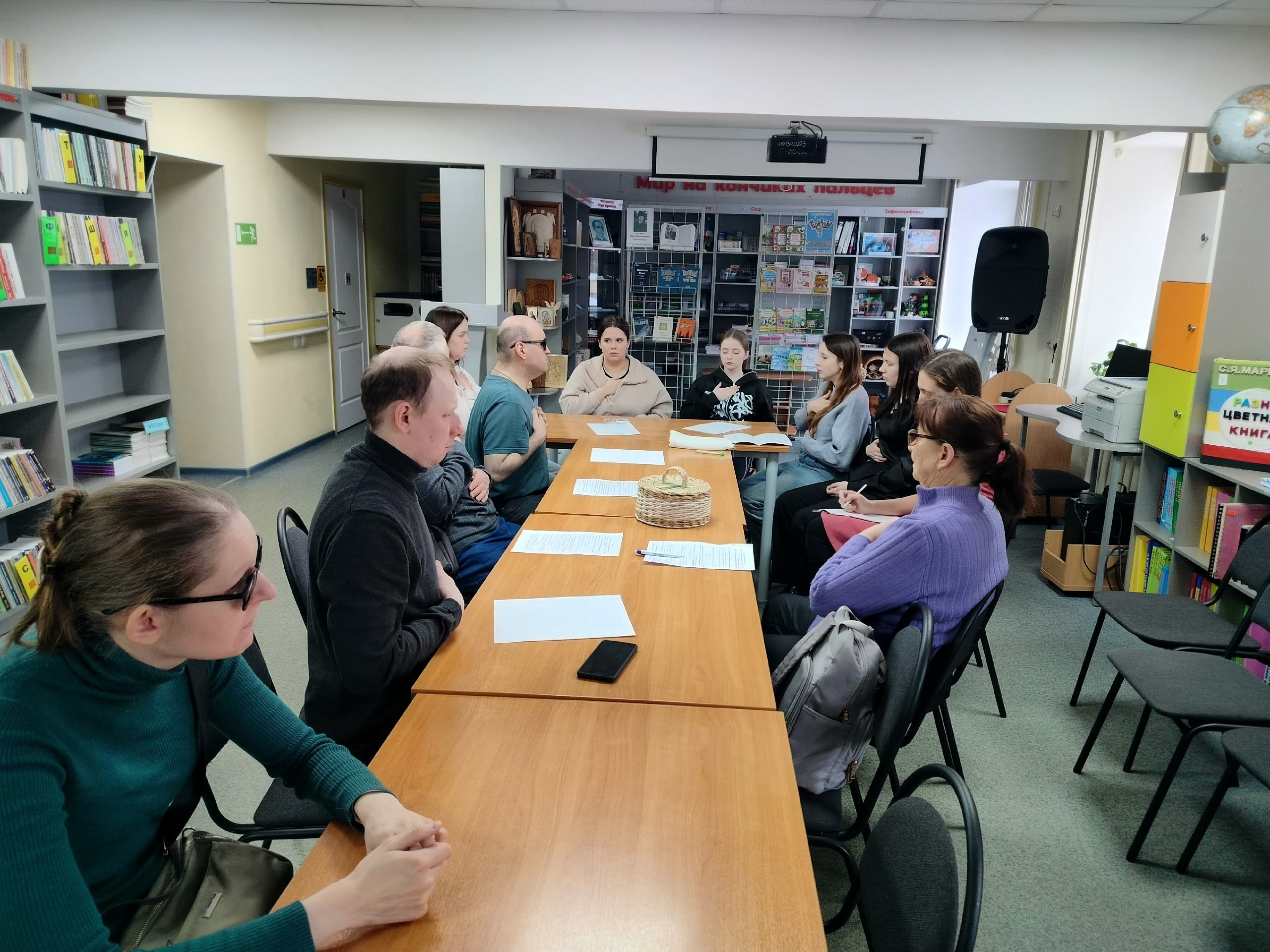Группа людей сидит за столами в библиотеке, некоторые смотрят на документы, вокруг книжные полки.