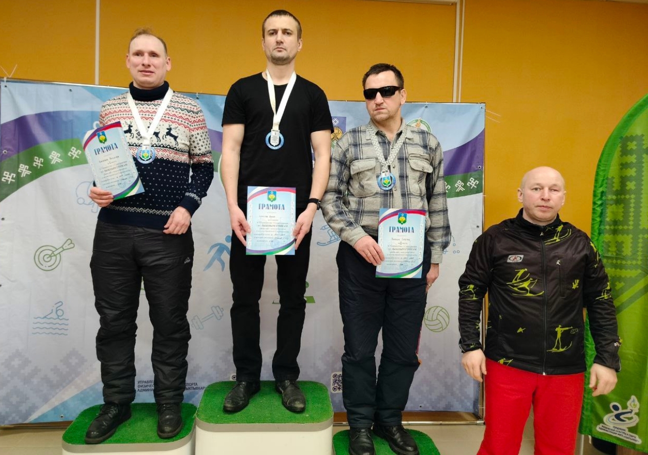 Трое мужчин с медалями и дипломами стоят на подиуме, справа мужчина в спортивной куртке.