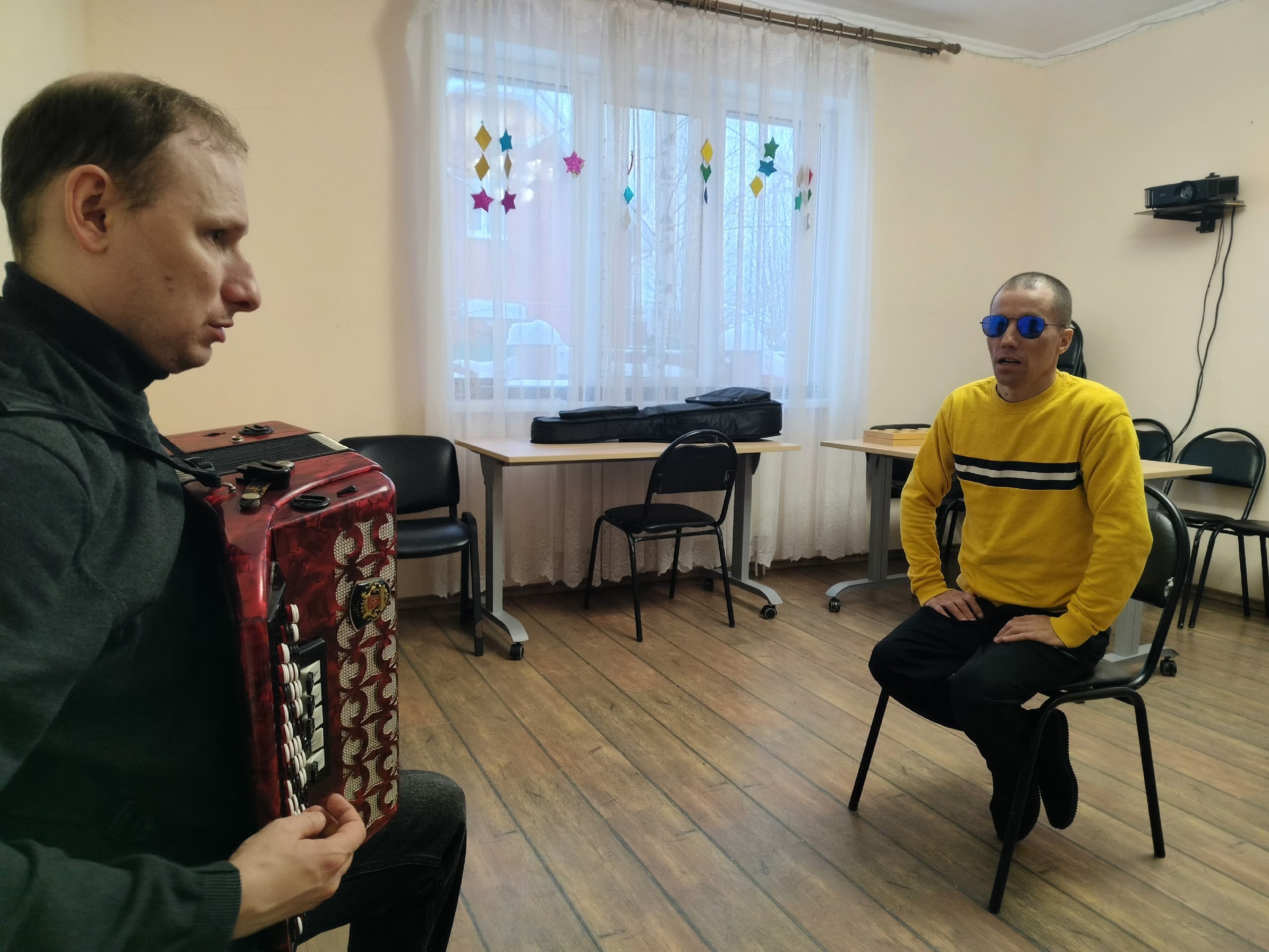 Мужчина в черном свитере играет на аккордеоне, а мужчина в желтом свитере с синими очками внимательно слушает.