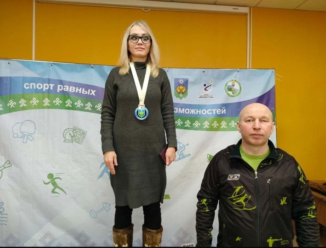 Женщина в очках и свитере с медалью на подиуме, рядом мужчина в спортивной куртке.