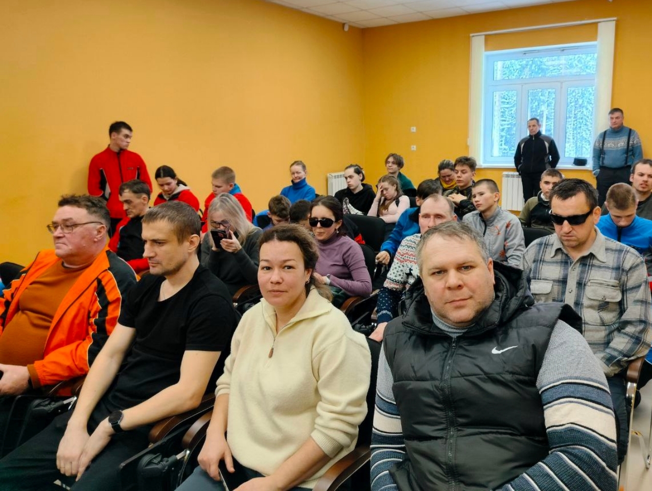 Группа людей сидит в зале, некоторые в темных очках, на фоне желтой стены и окна.