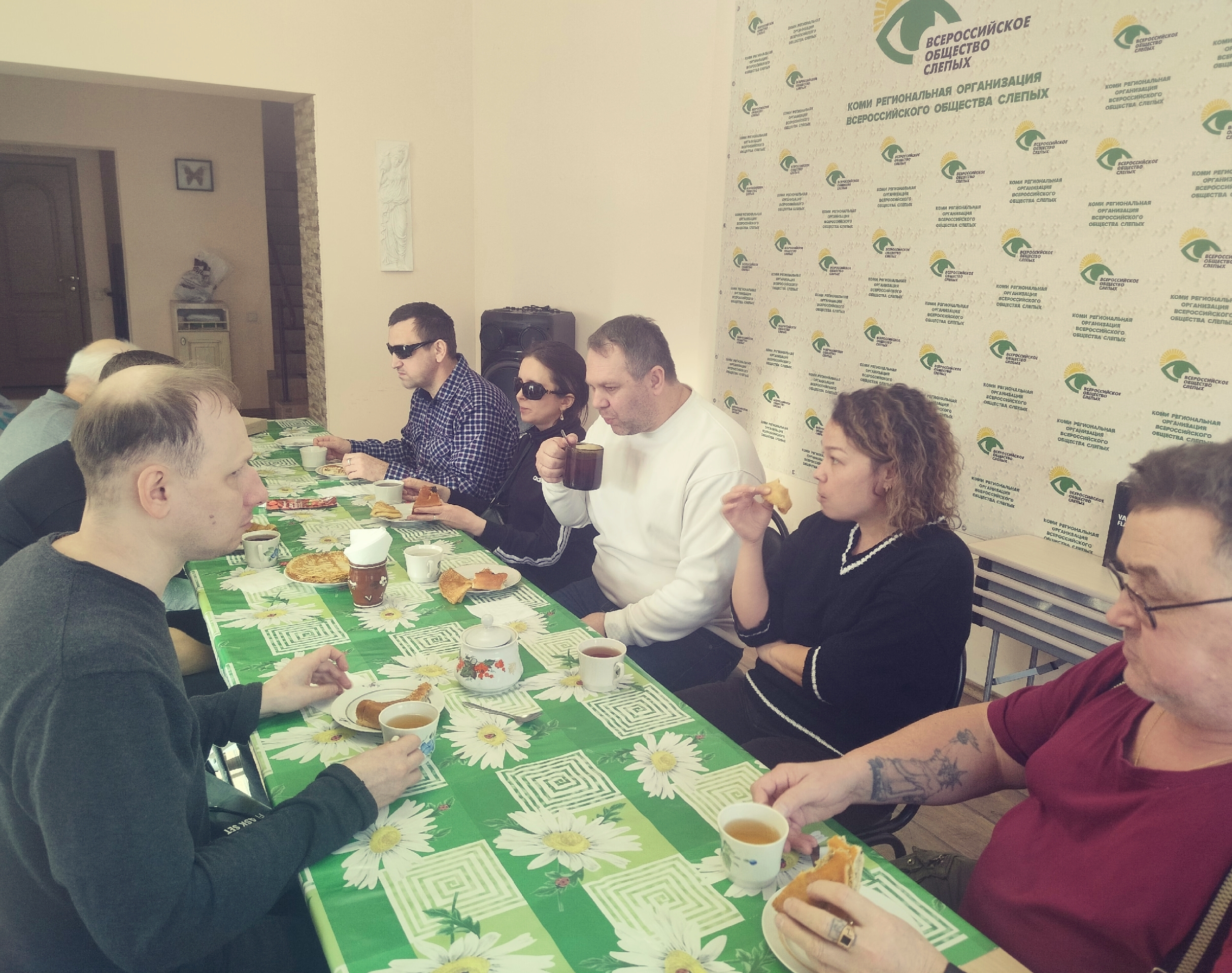 Группа мужчин и женщин пьют чай с угощениями за столом, накрытым зеленой скатертью, на фоне баннера Всероссийского общества слепых.