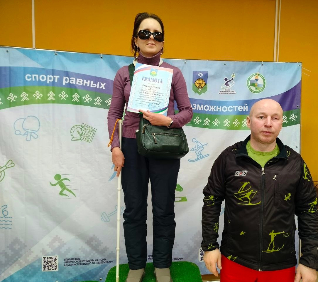 Женщина в темных очках с дипломом и тростью на подиуме, рядом мужчина в спортивной куртке.