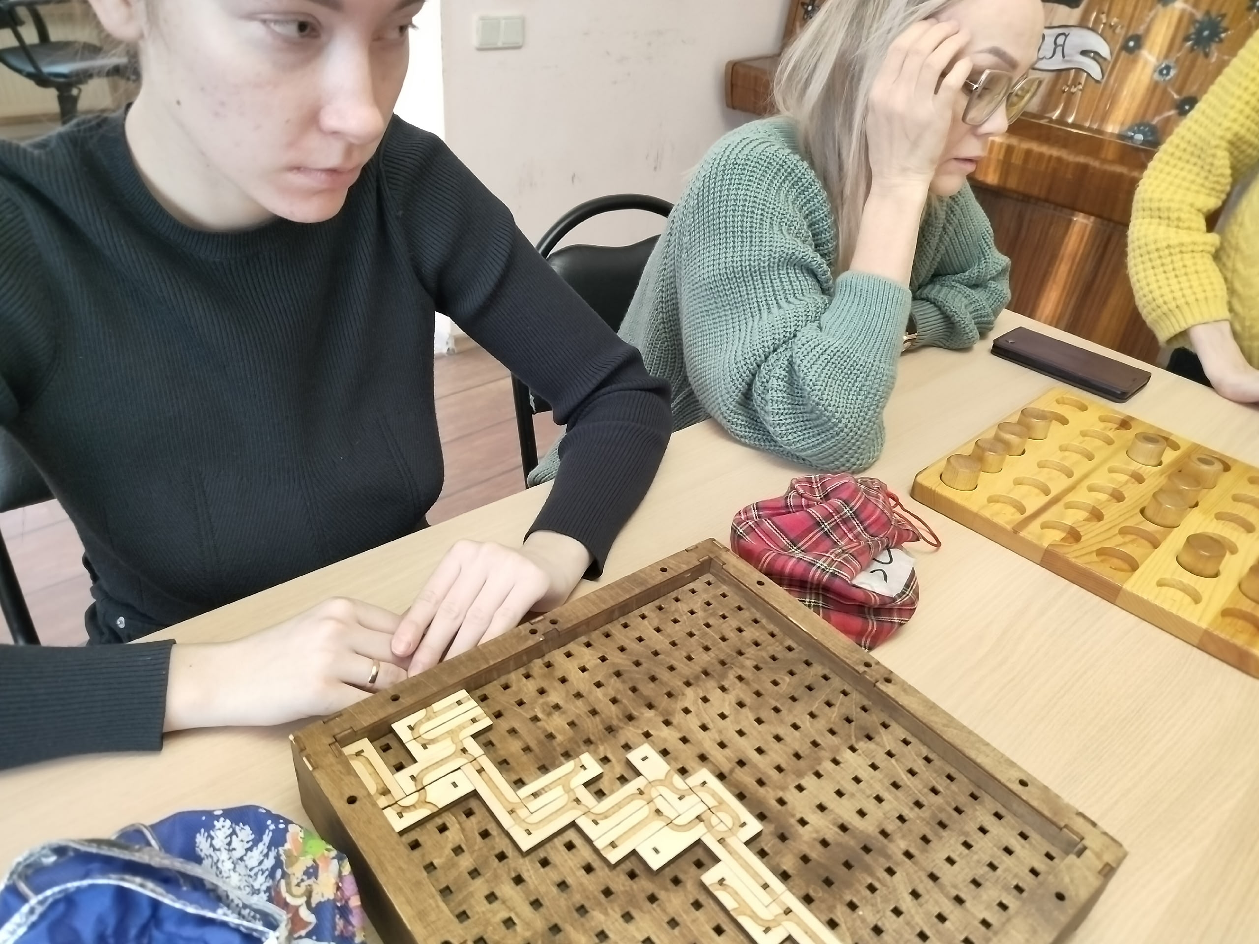 Две женщины играют в настольные игры: одна сосредоточена на деревянной головоломке, другая смотрит на игровое поле.