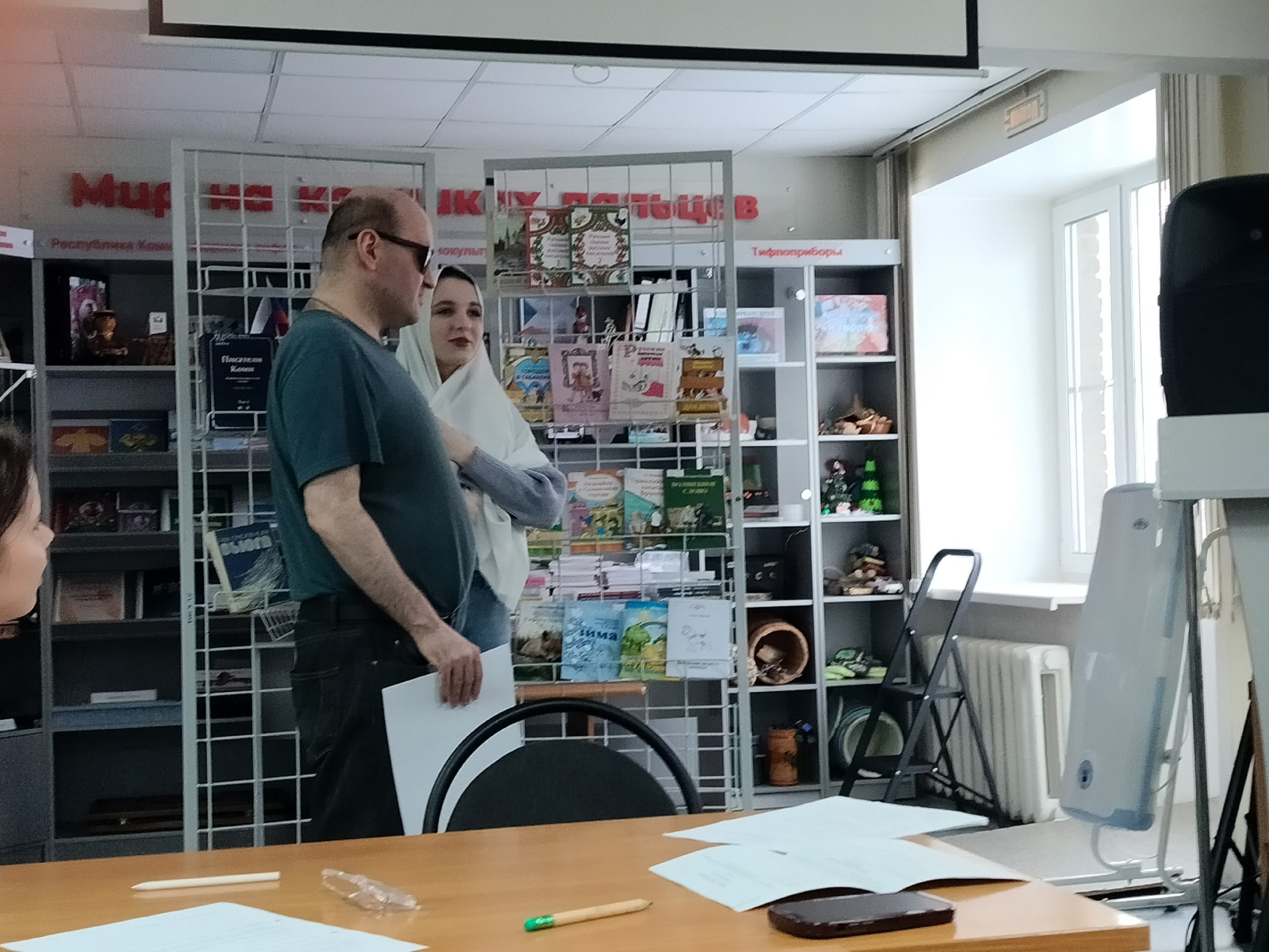 Мужчина в очках и женщина в платке стоят в библиотеке у книжных стеллажей, на столе лежат бумаги.