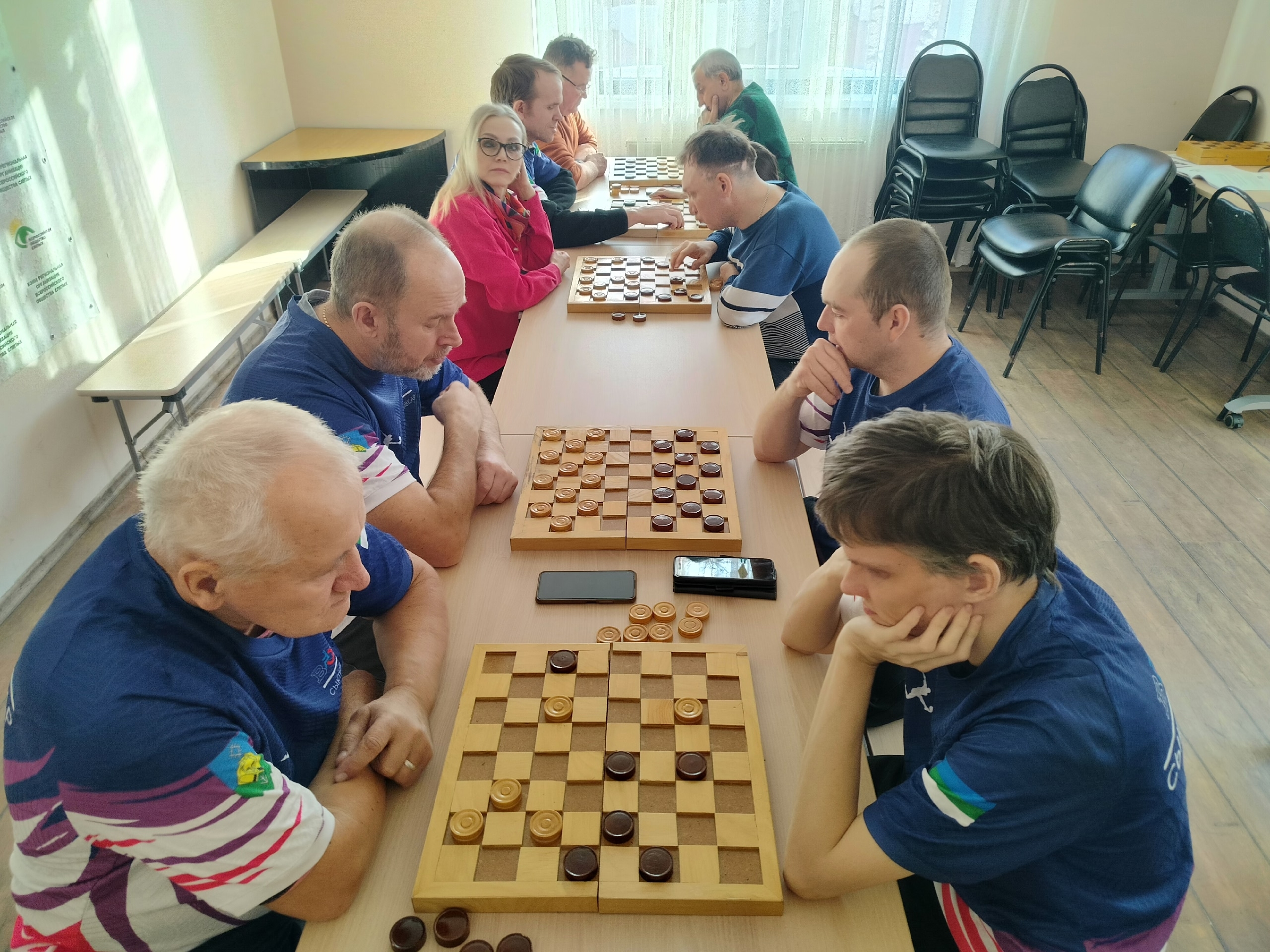 Общий вид на турнир по шашкам, участники сидят за столами, увлеченные игрой и обдумывающие ходы.