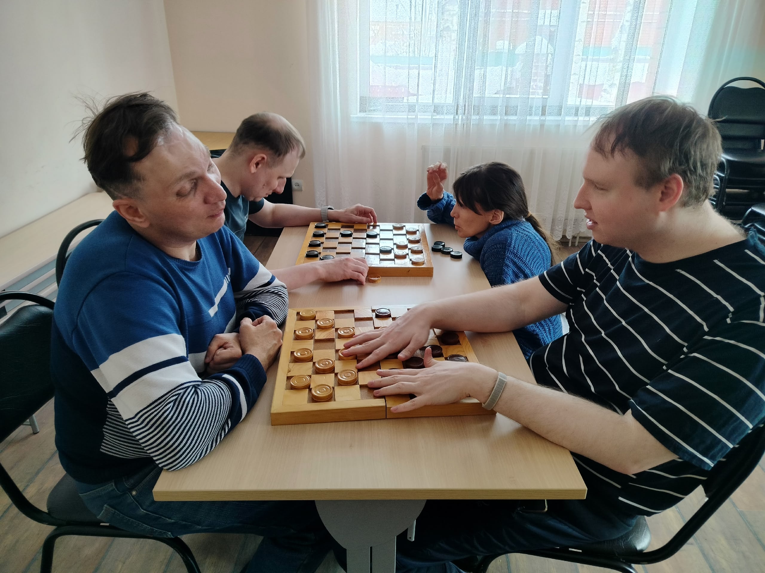 Четыре человека, включая троих мужчин и одну женщину, сидят за столом и играют в шашки на двух досках в помещении.