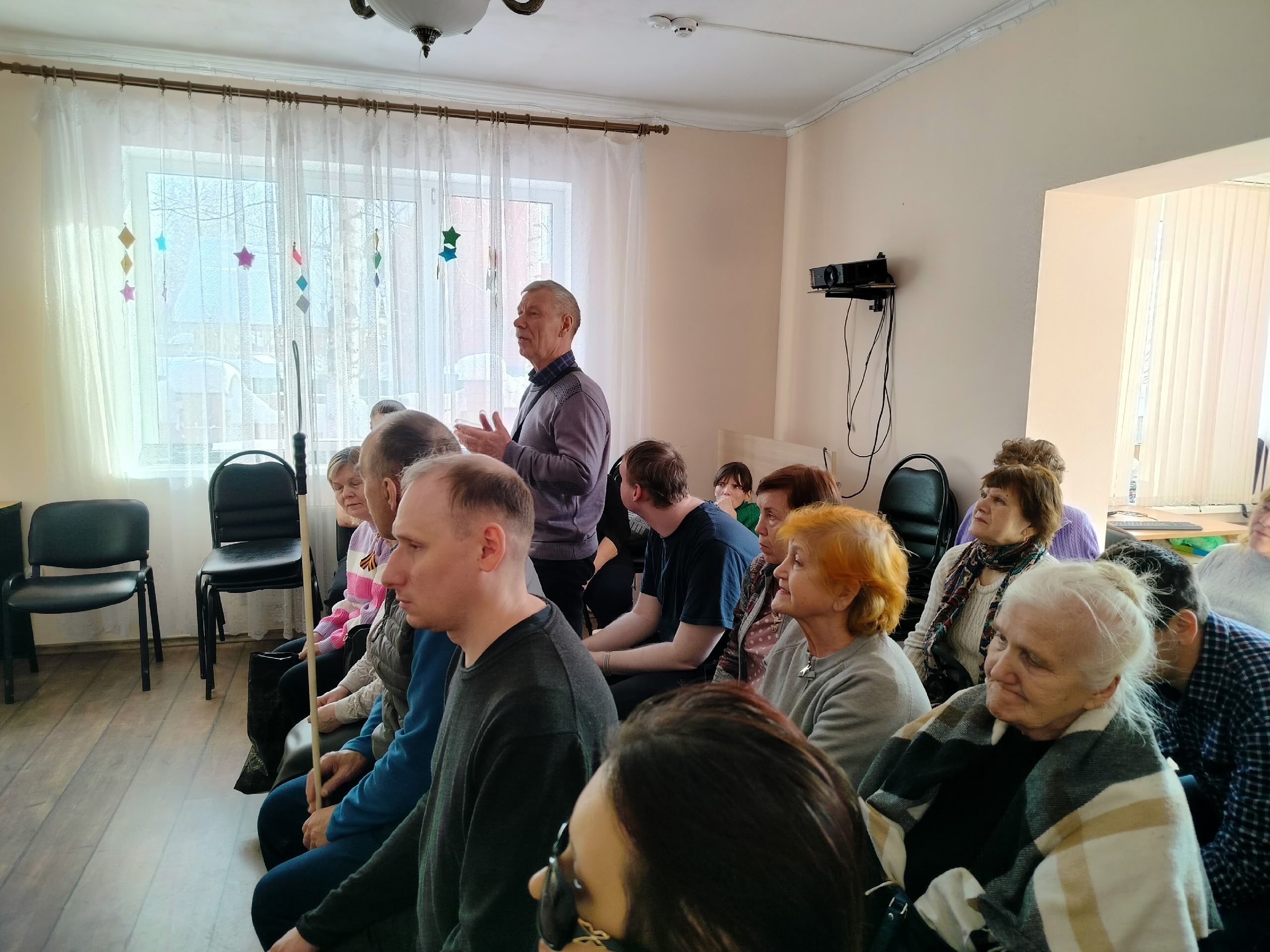 Мужчина в сиреневом свитере стоит и говорит, обращаясь к сидящей аудитории в светлом помещении.