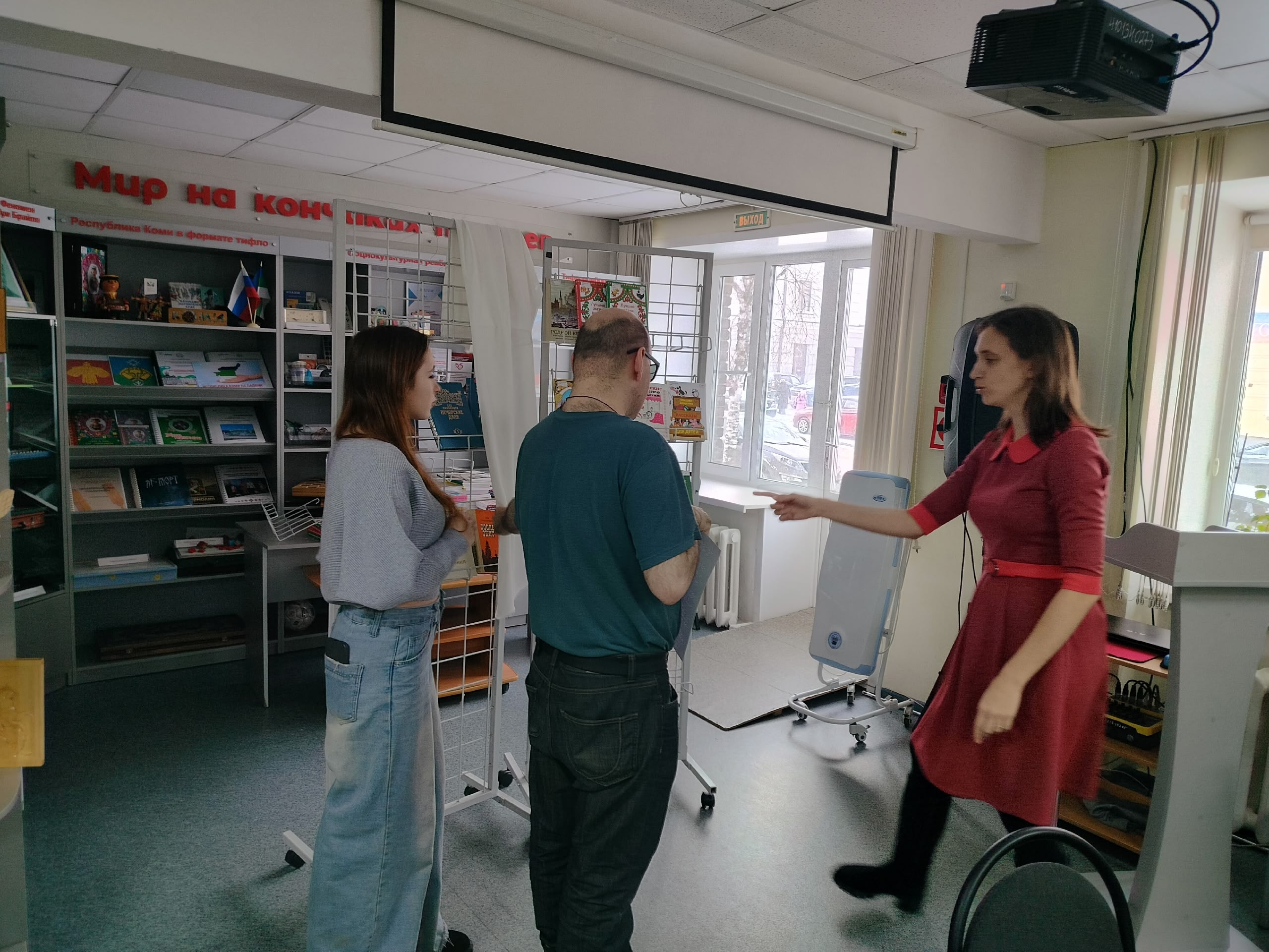 Три человека стоят в библиотеке: женщина в красном платье жестикулирует, обращаясь к мужчине, рядом стоит девушка.