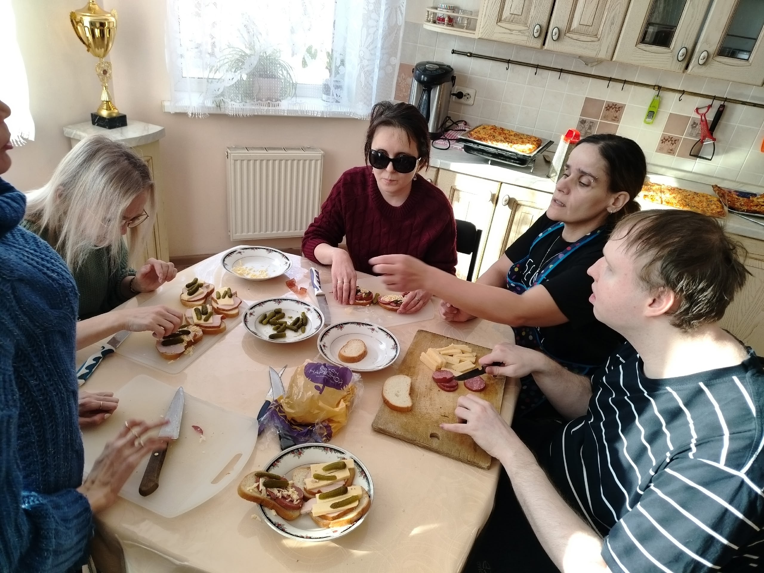 Группа людей, включая женщину в темных очках, готовит бутерброды с сыром, колбасой и корнишонами на кухне.
