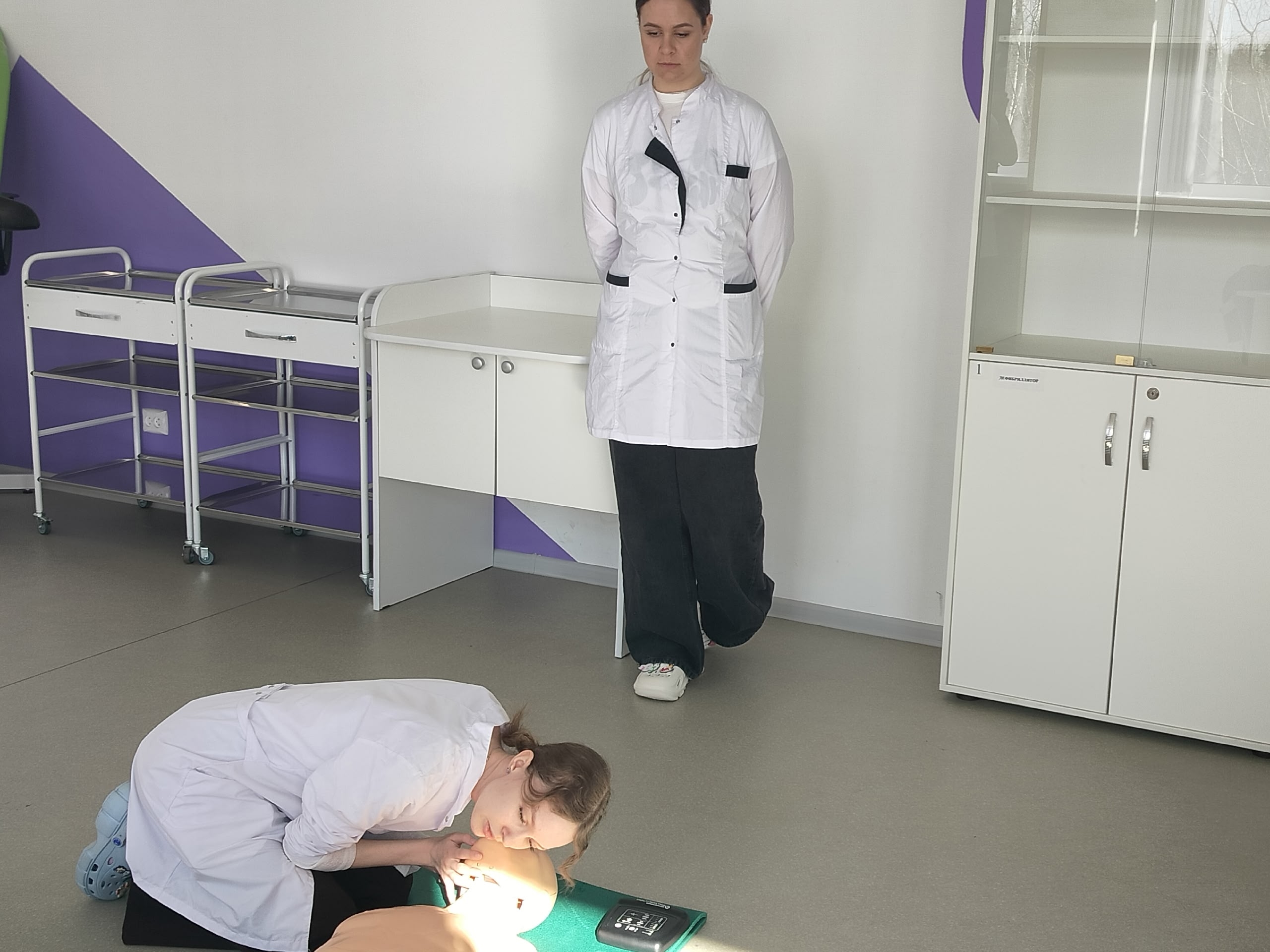 Женщина в халате проводит реанимацию на учебном манекене.