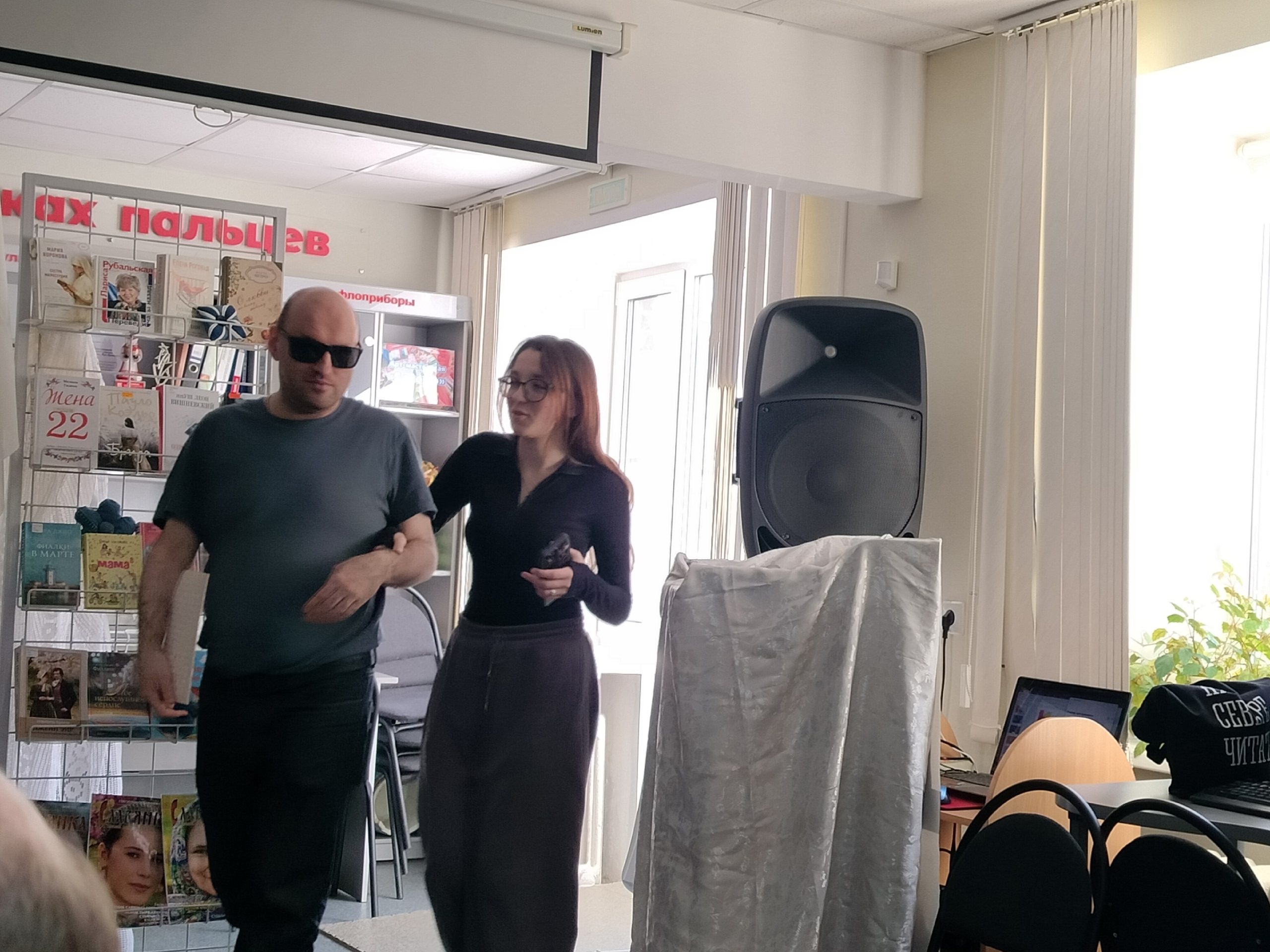 Мужчина в темных очках и женщина в очках идут по помещению библиотеки во время репетиции.