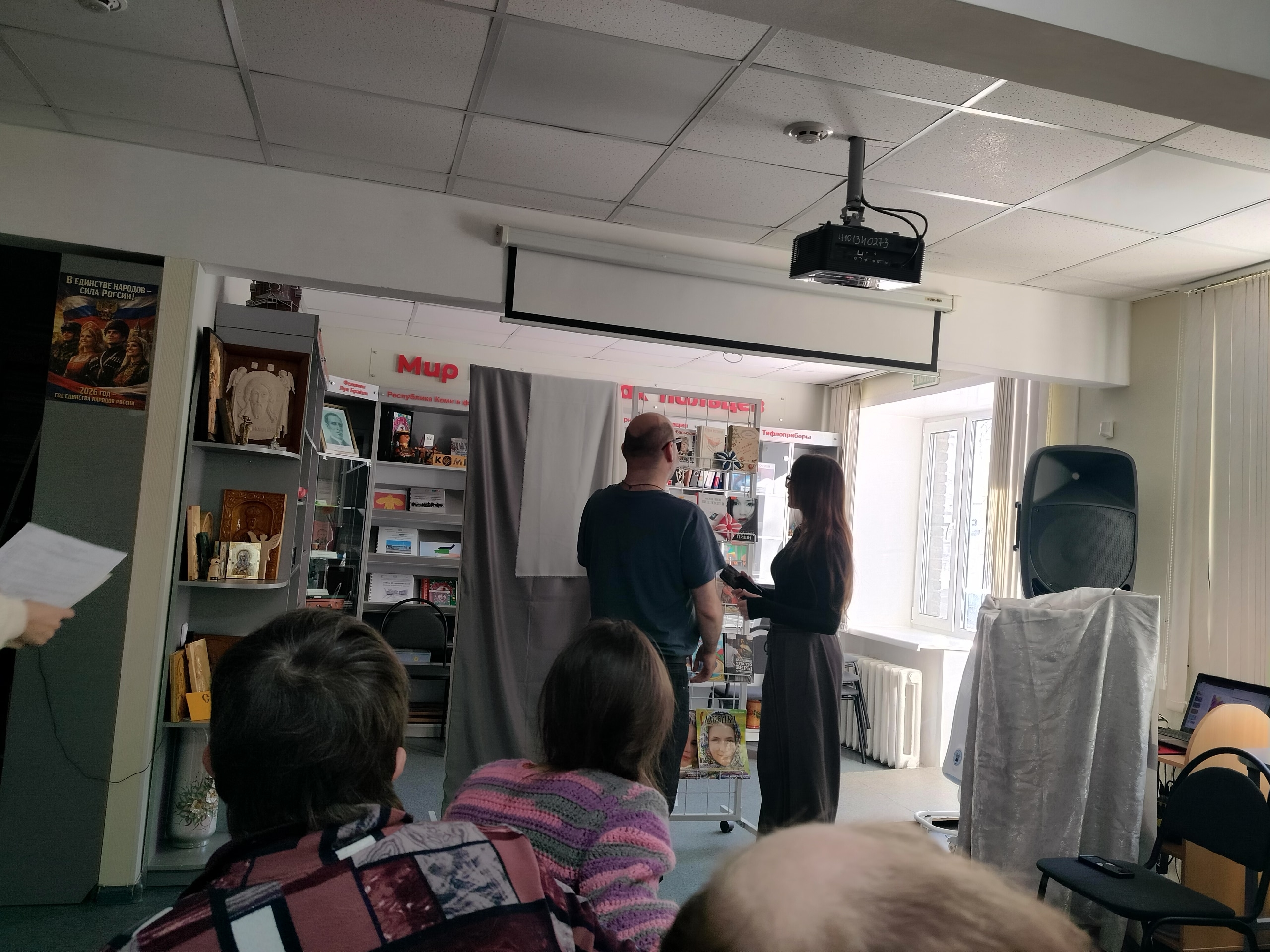Репетиция культурного мероприятия в библиотеке, где мужчина и женщина стоят перед аудиторией.