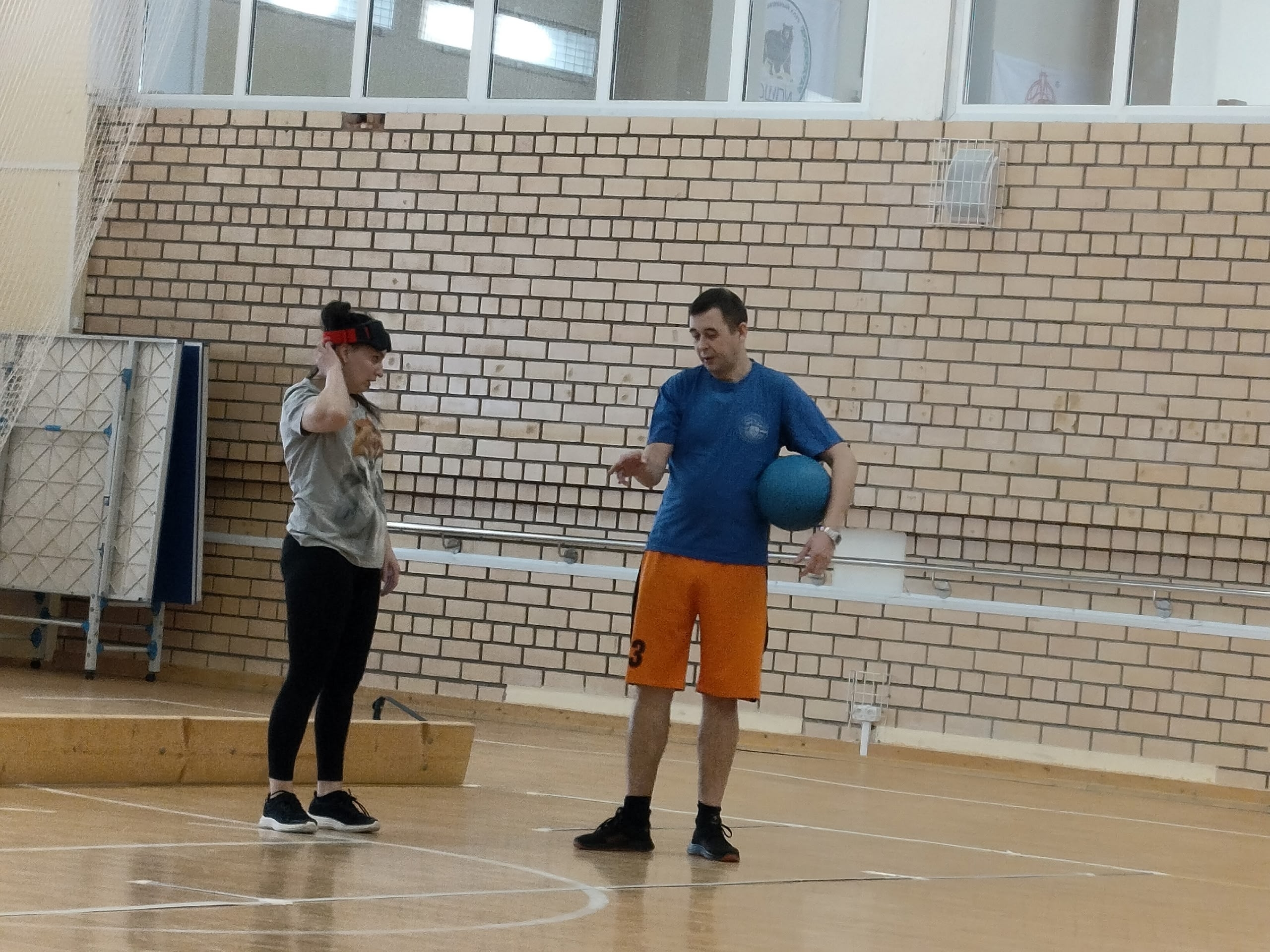 Мужчина в синей футболке и оранжевых шортах держит синий мяч, разговаривает с женщиной.