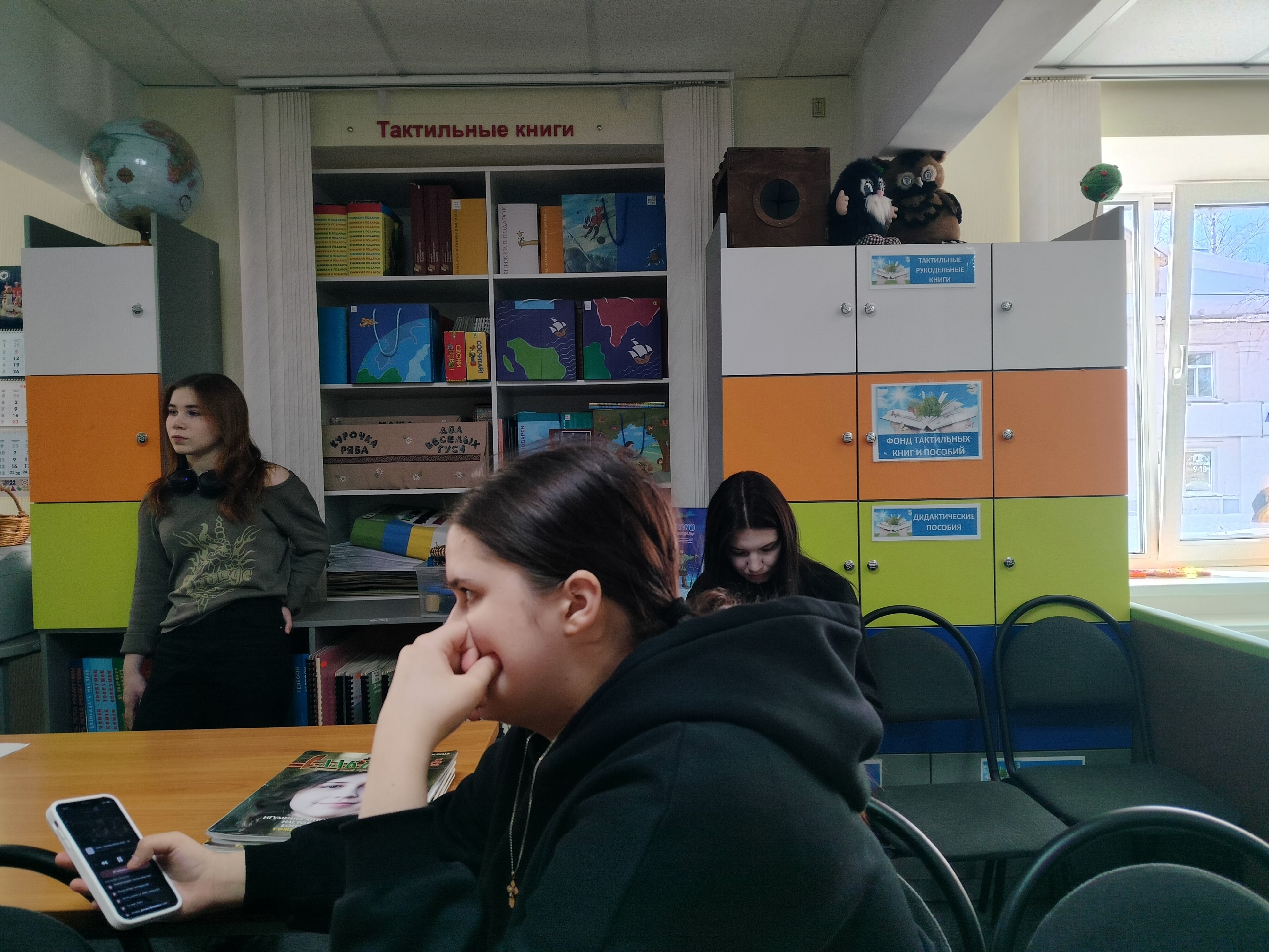 Девушки сидят и стоят в помещении библиотеки с тактильными книгами на полках и разноцветными шкафчиками.