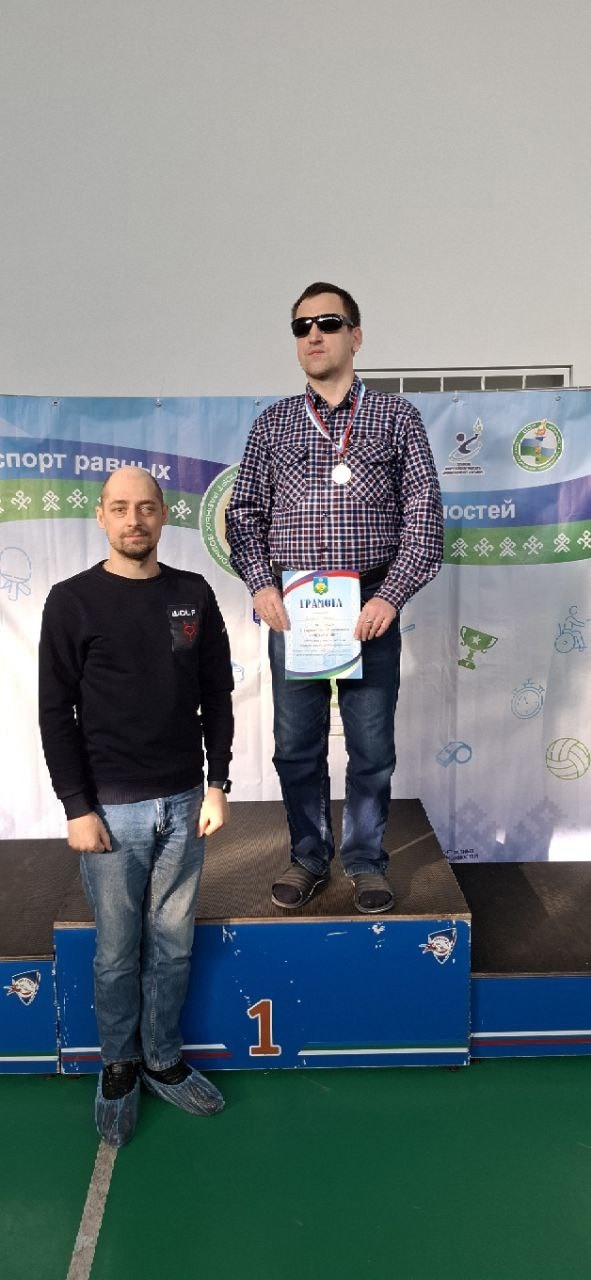 Двое мужчин на пьедестале, один в темных очках получает медаль и грамоту.