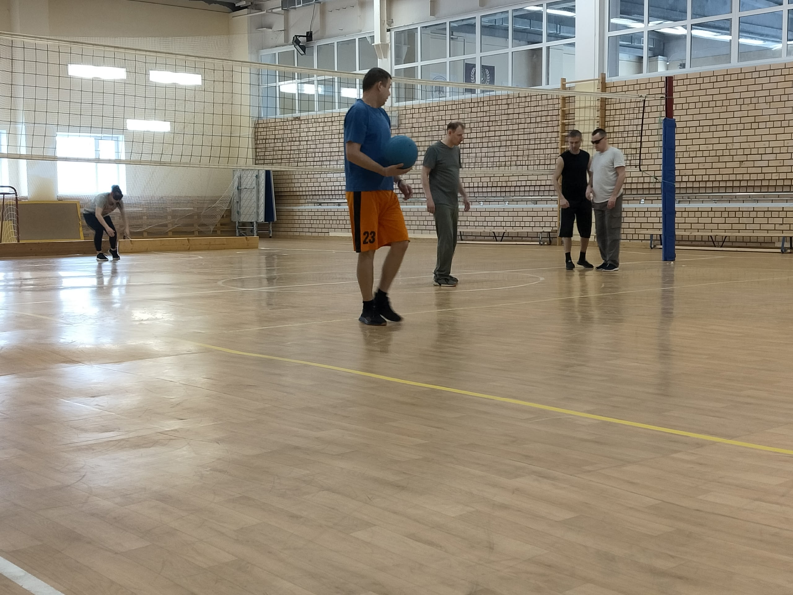 Мужчина в синей футболке и оранжевых шортах идет с синим мячом по спортивной площадке.