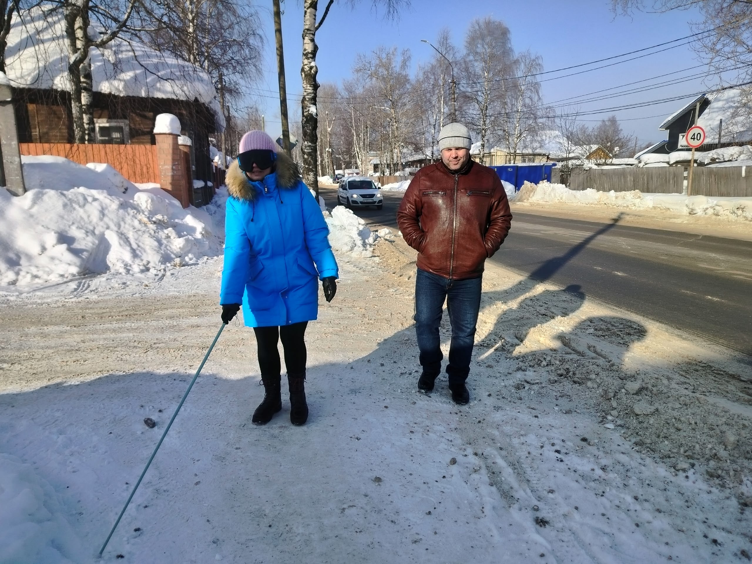 Женщина в синей куртке и розовой шапке с повязкой на глазах идет по заснеженной улице, опираясь на белую трость, рядом идет мужчина.