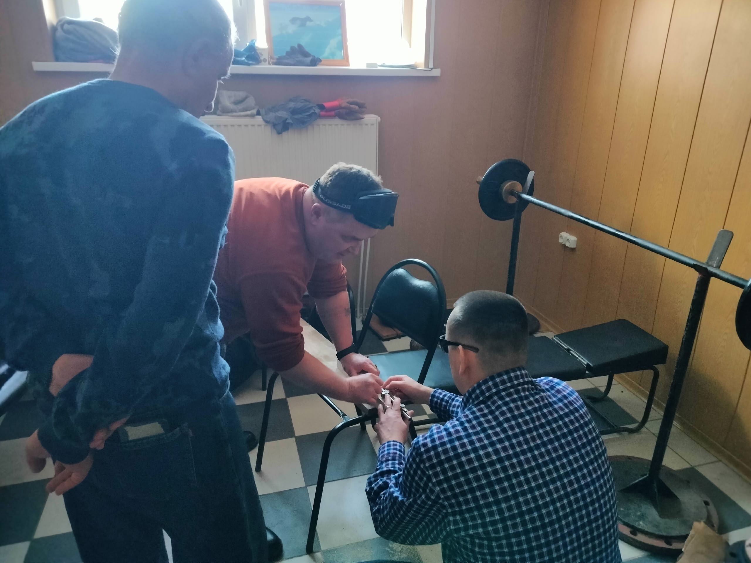 Трое мужчин в помещении, один из них в повязке на глазах, работает с деревянным бруском на подставке.