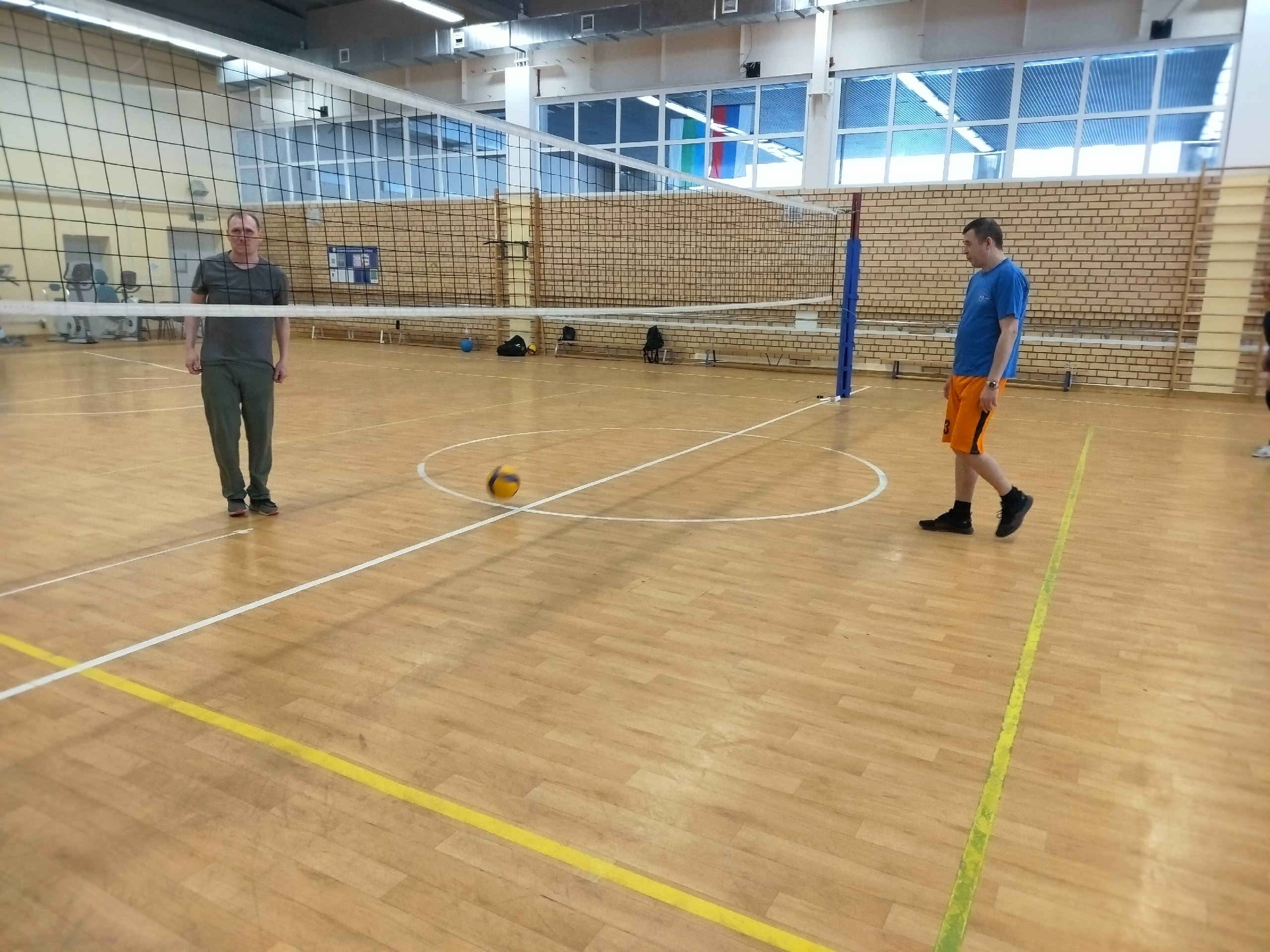 Два мужчины на волейбольной площадке, один в синей футболке и оранжевых шортах, с мячом на полу.
