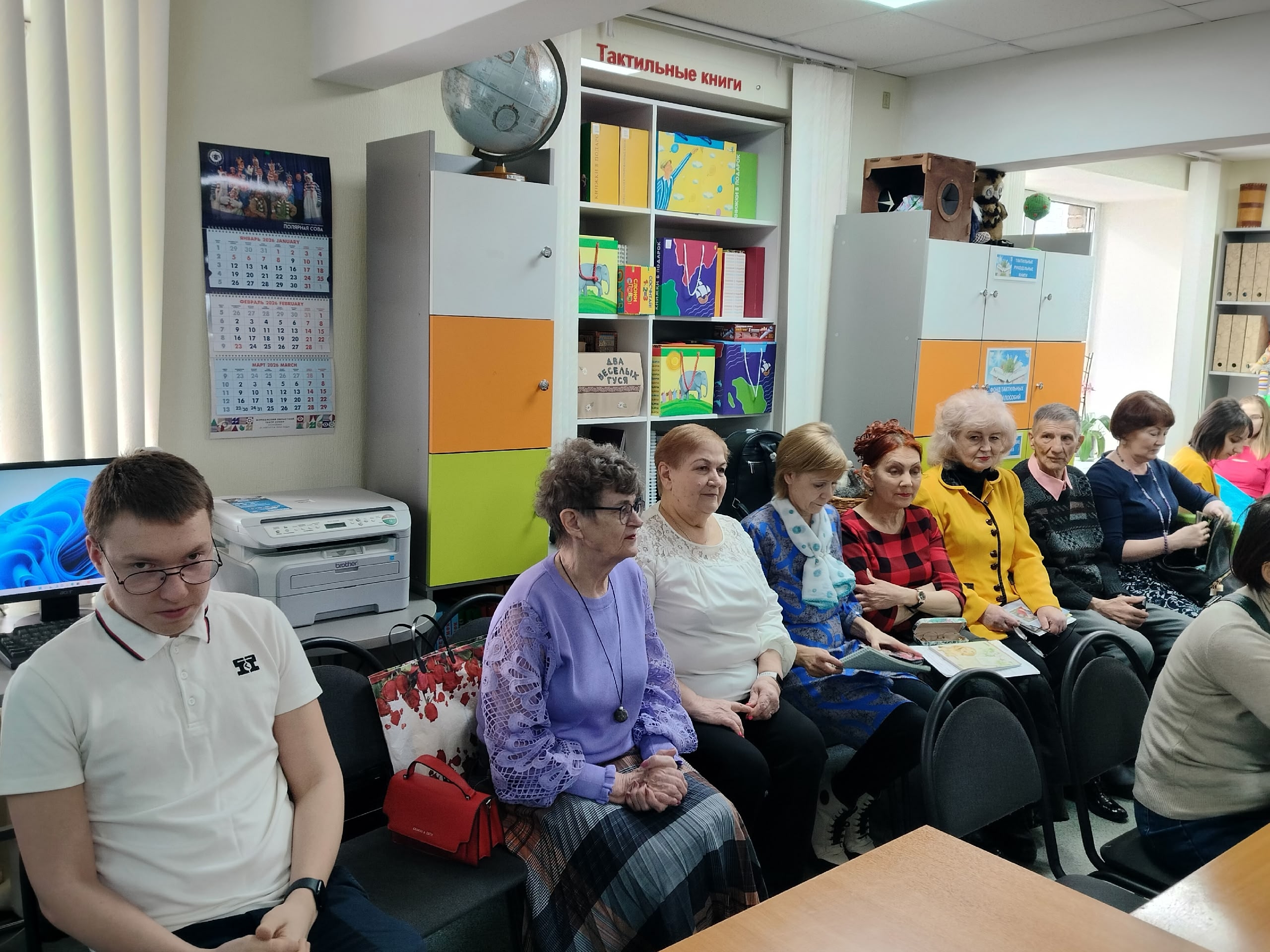 Группа людей сидит в библиотеке, слушая выступление.