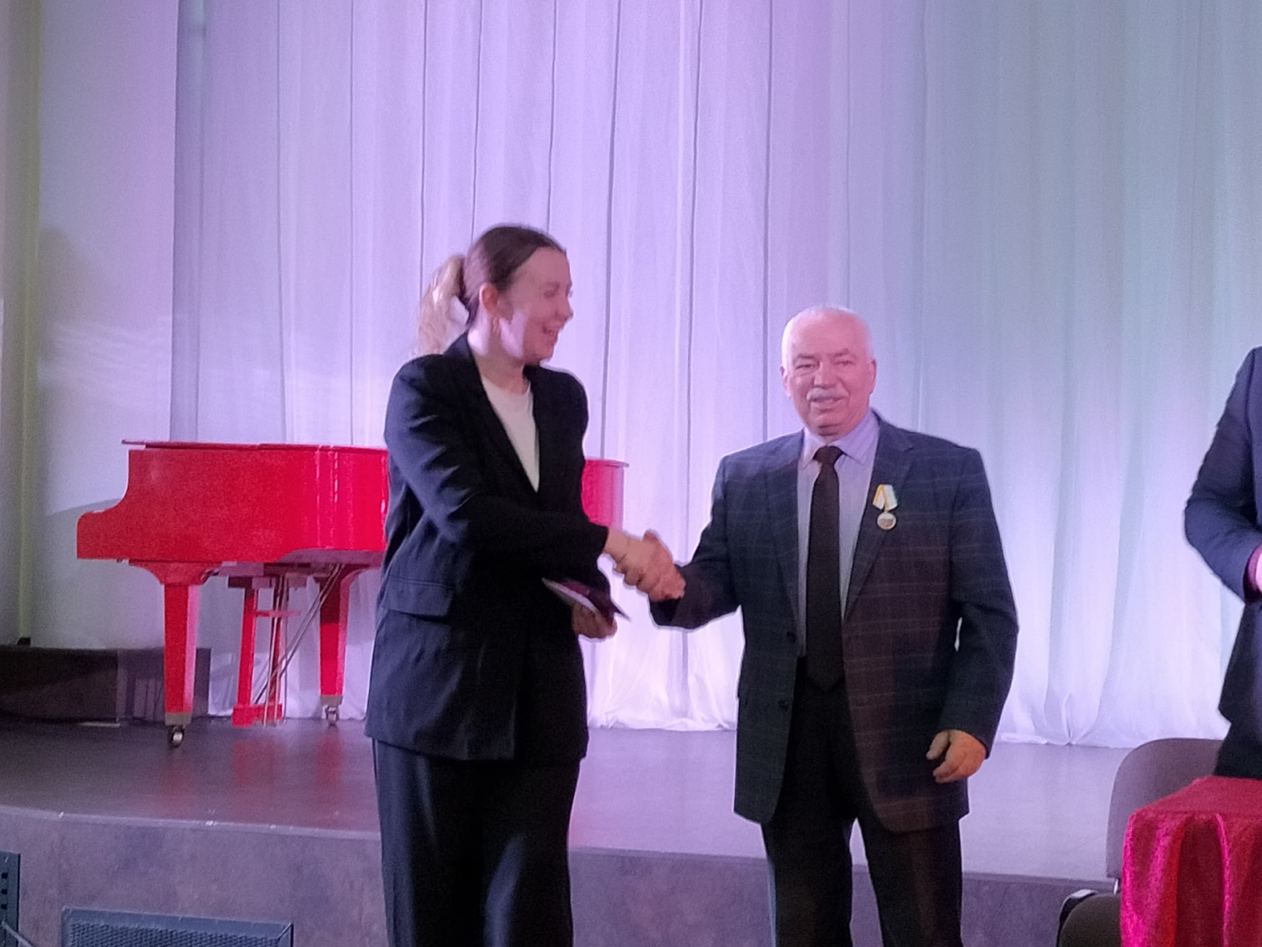 Женщина в темном костюме пожимает руку мужчине в клетчатом пиджаке с медалью на сцене.