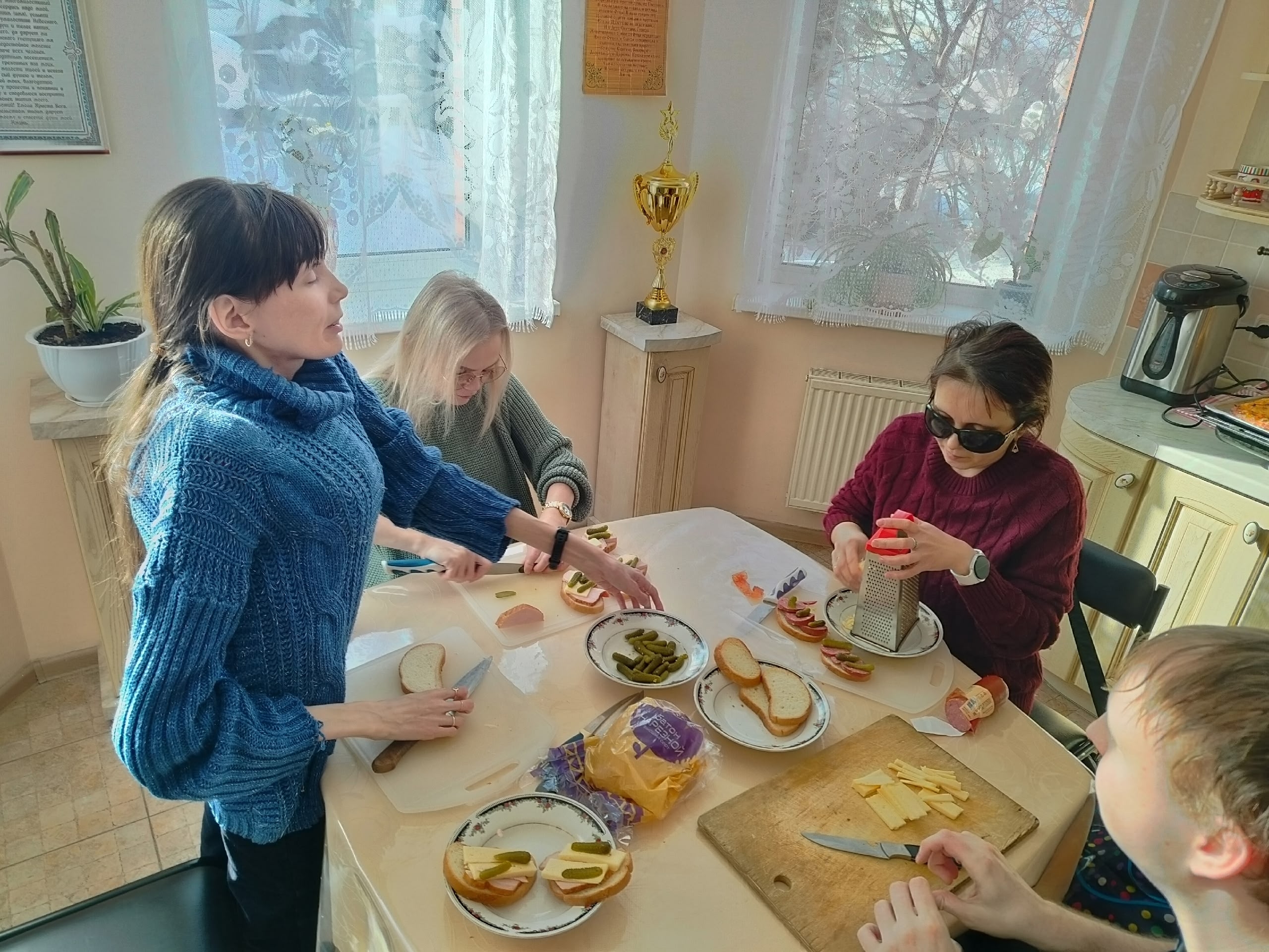 Женщины и мужчина готовят бутерброды на кухне, одна из них в темных очках трет сыр на терке.