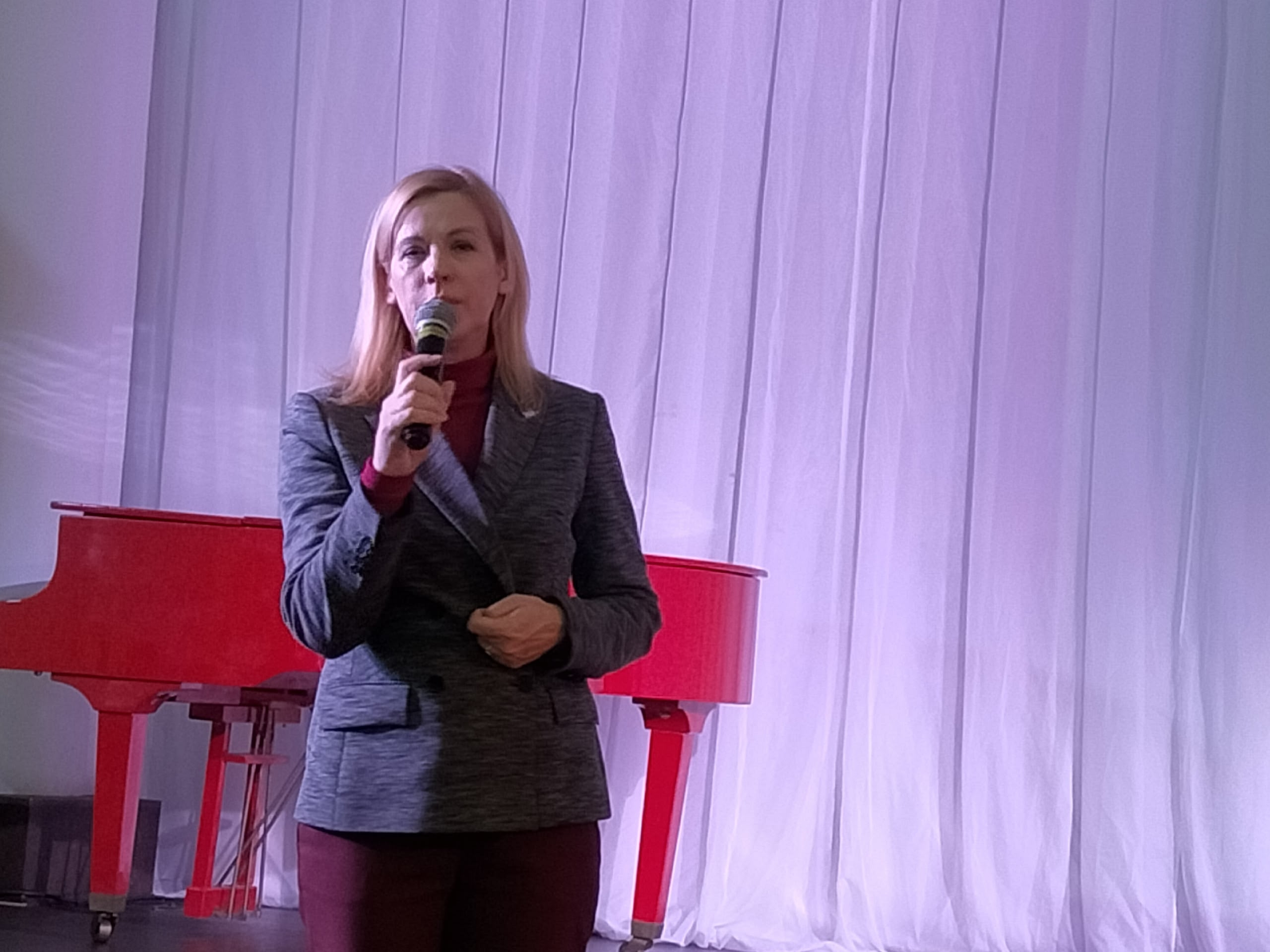 Женщина в сером пиджаке и бордовой кофте стоит на сцене, держа микрофон.