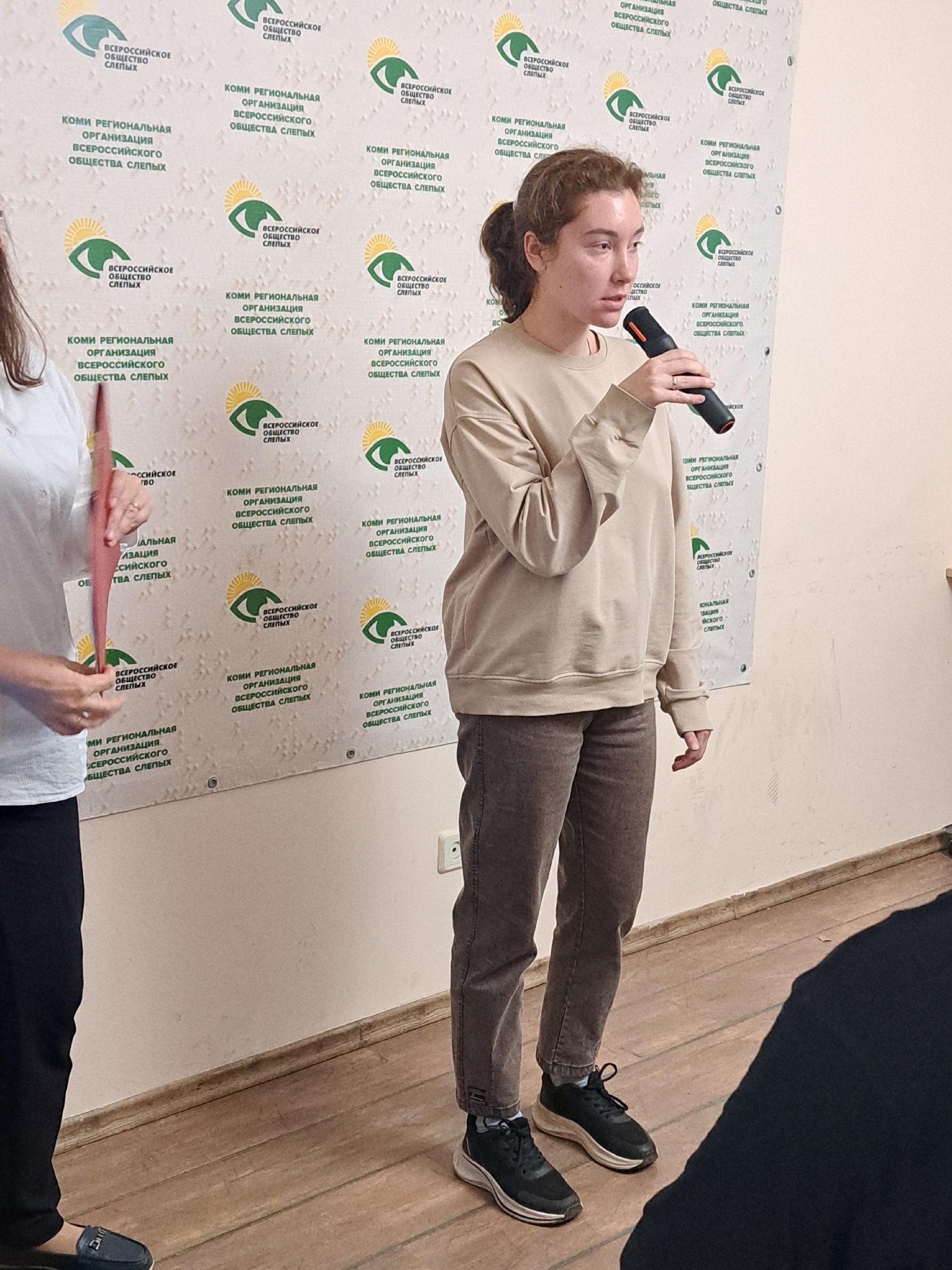 Девушка в бежевой кофте держит микрофон и выступает