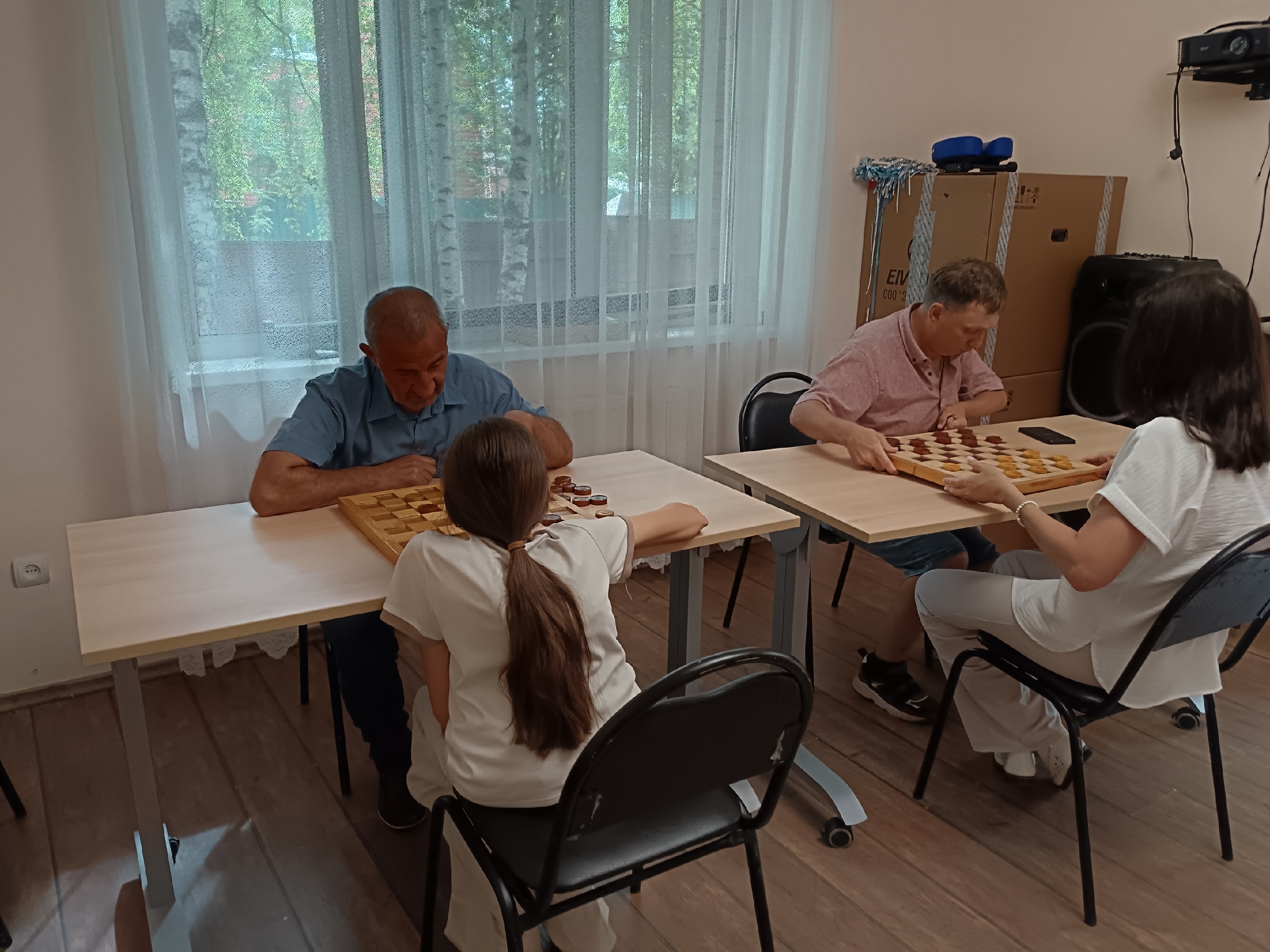 Две пары игроков сидят за столами и играют в шашки в светлой комнате