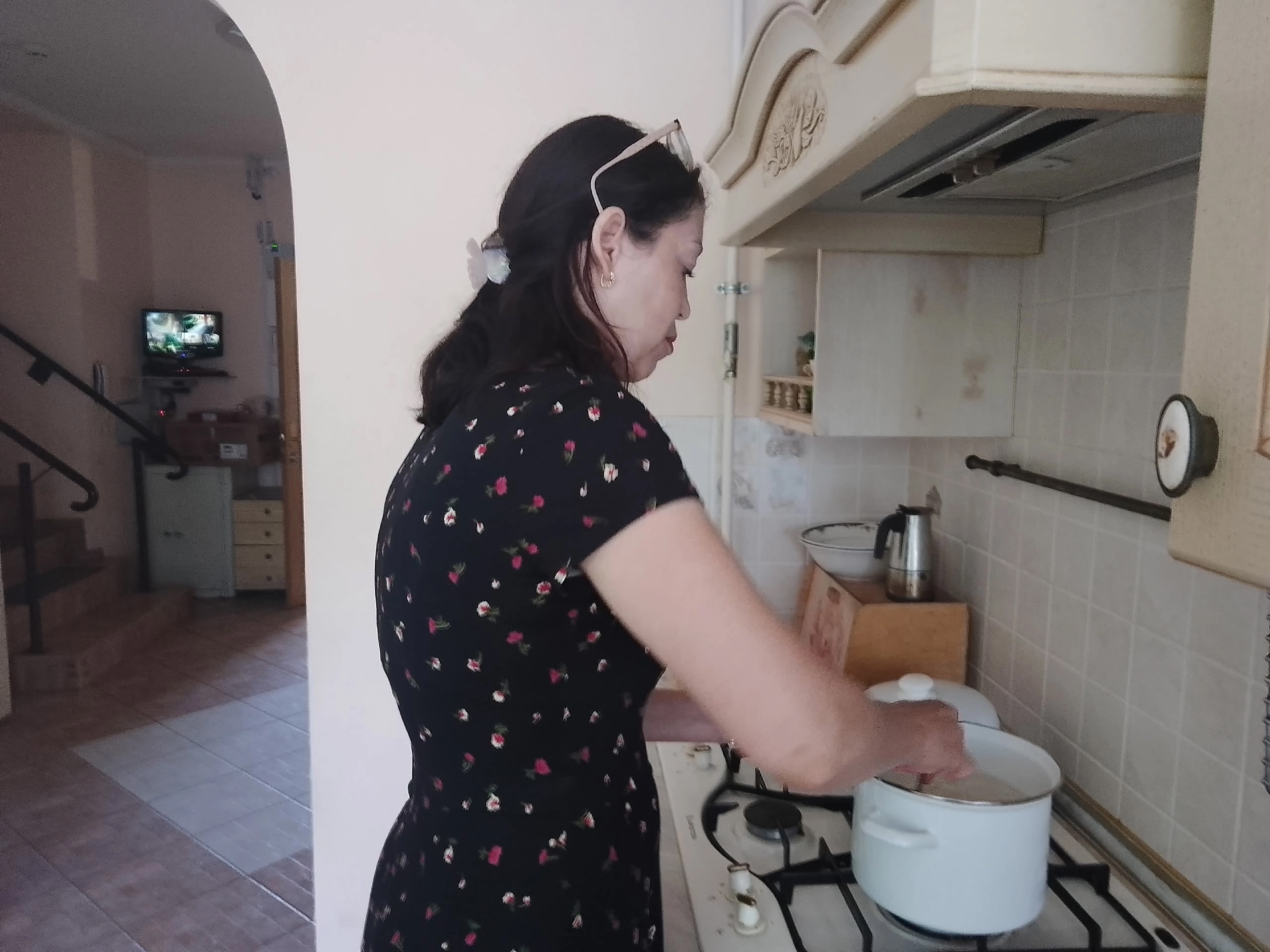 Женщина в темном платье в цветочек помешивает блюдо в белой кастрюле на газовой плите