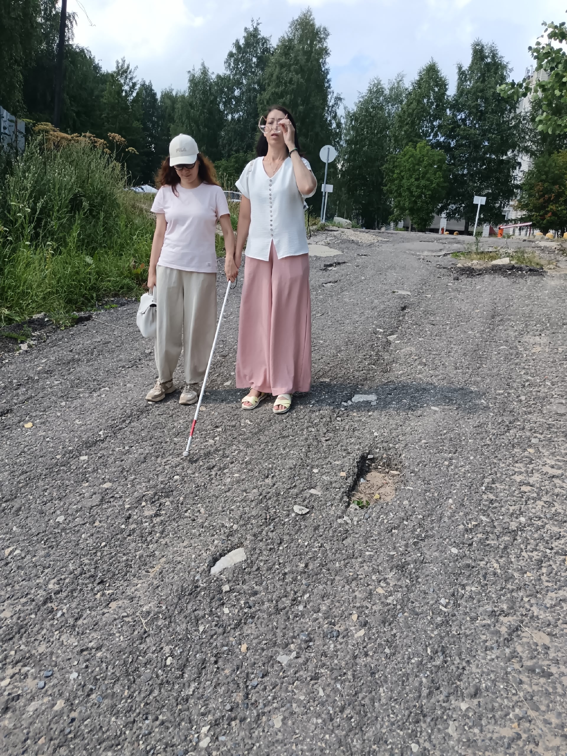 Две женщины идут по разбитой асфальтовой дороге, одна из них использует тактильную трость