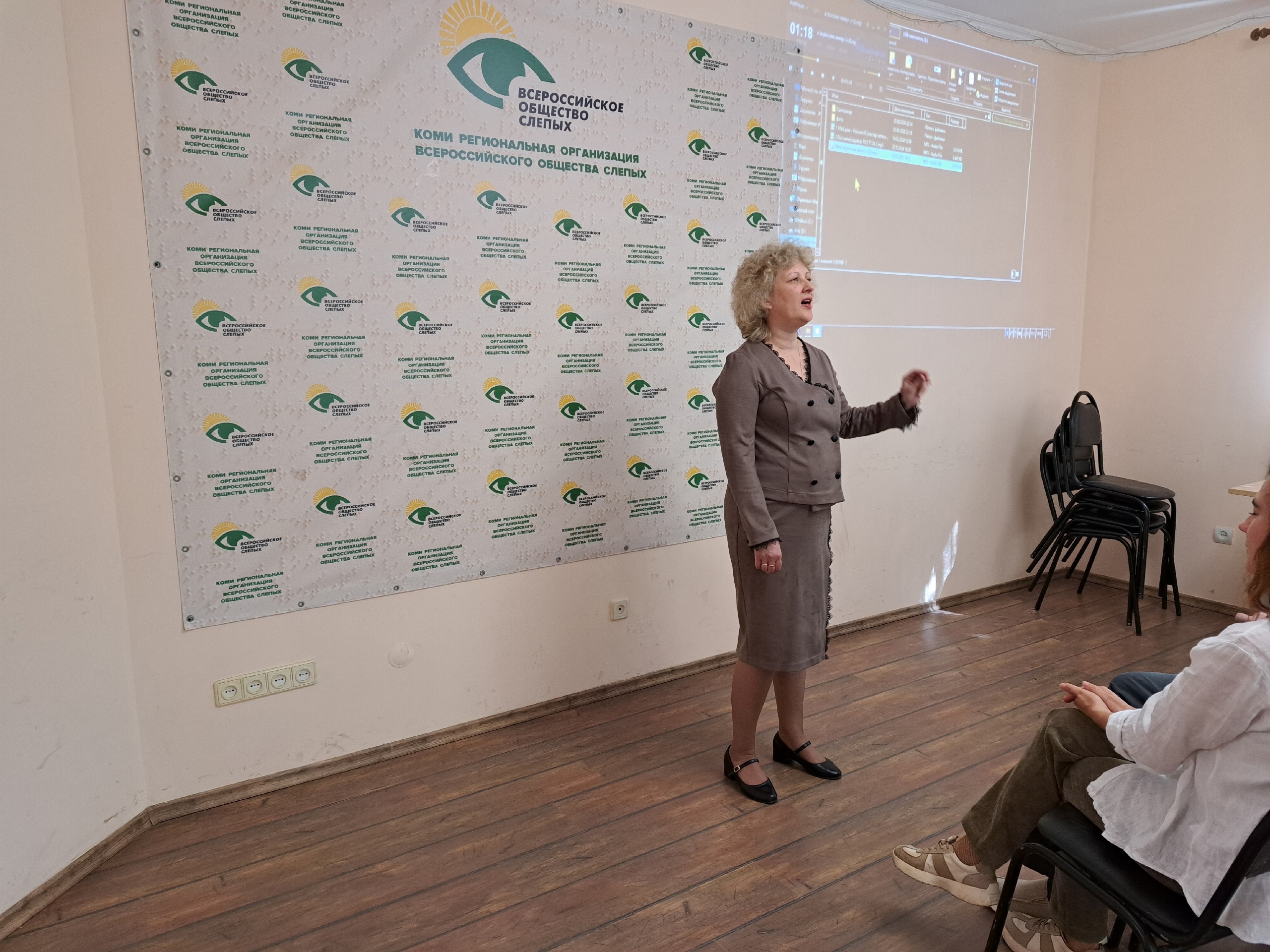 Женщина в сером костюме выступает перед слушателями на фоне баннера Общества слепых