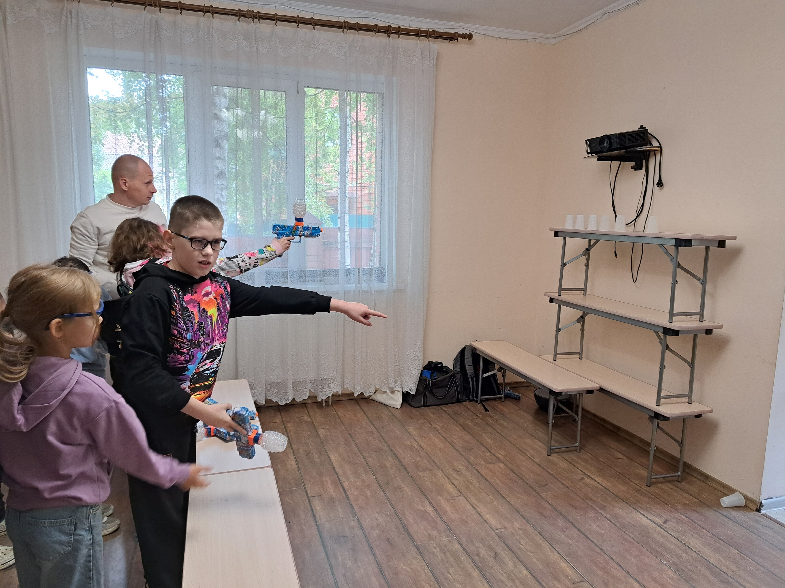Мальчик в очках целится из игрушечного пистолета в пластиковые стаканчики