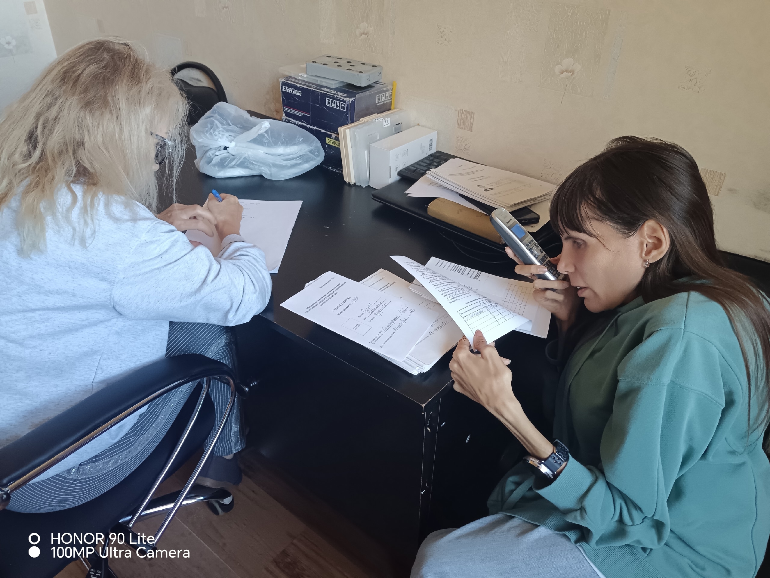 Две женщины за столом работают с документами и совершают телефонные звонки